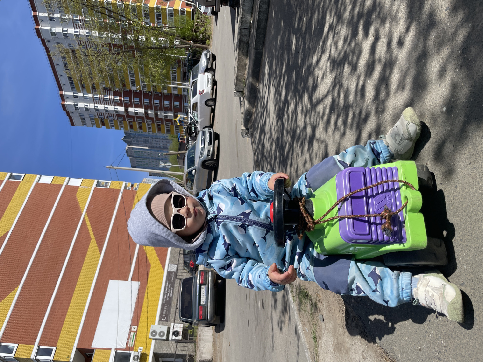 A child wearing a blue patterned jacket, grey hooded sweatshirt, sunglasses, and grey sneakers is sitting on a green and purple toy on an asphalt road. The child faces the camera with a neutral expression. In the background, there are residential buildings with yellow and brown striped walls and white apartments, along with parked cars. The scene is set in Izhevsk, Russia, during daylight with shadows cast by trees.
