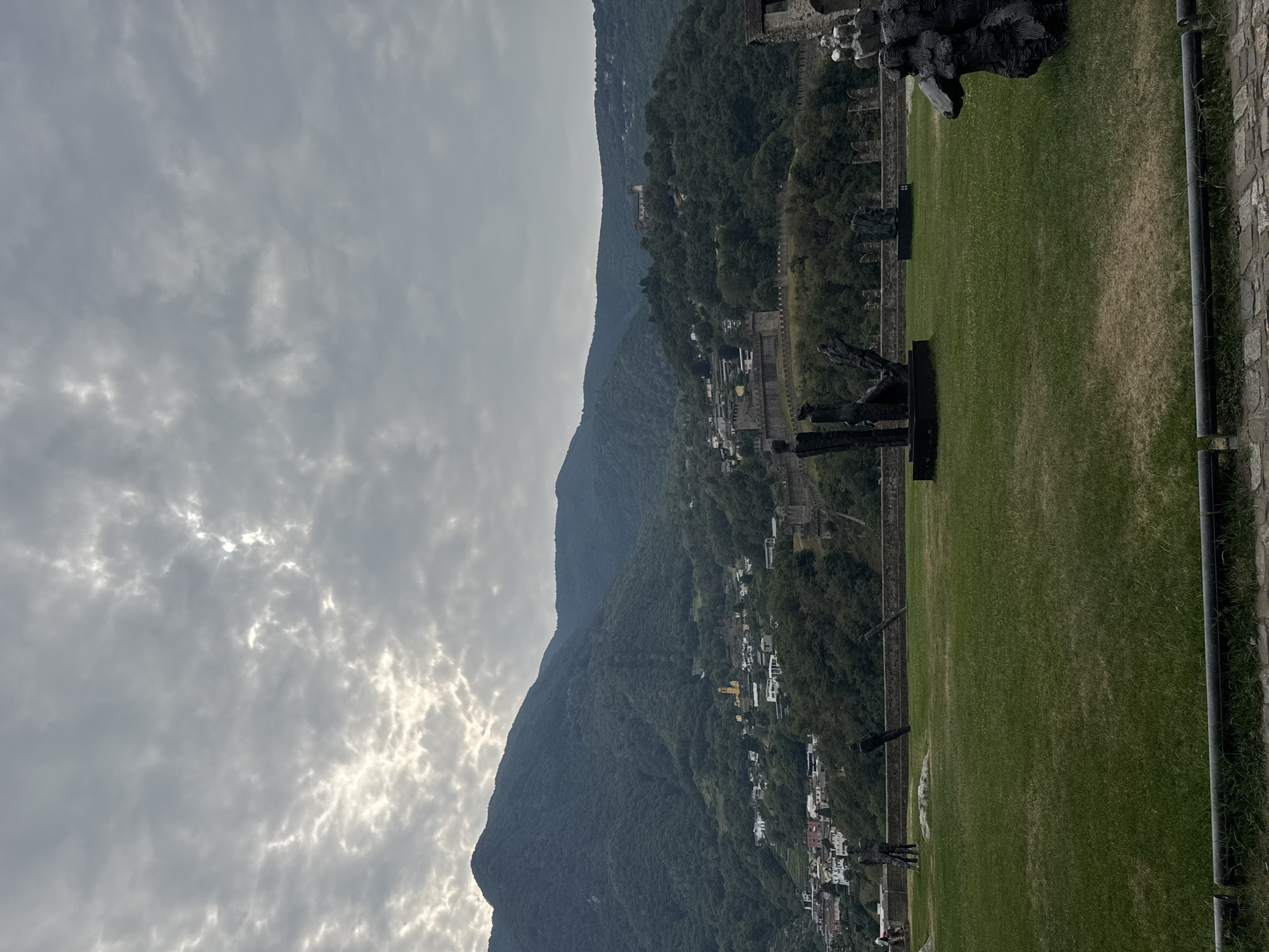 In Bellinzona, Switzerland, a scenic overlook frames a valley dotted with buildings and ancient fortifications. The foreground features a meticulously manicured lawn, adorned with several abstract, dark-colored sculptures. Two prominent human-like figures stand near the center, their forms elongated and somewhat distorted. To the right, a larger, more organic sculpture depicts a seated figure. The middle ground reveals a stone wall, likely part of a historical fortress, stretching across the landscape. Beyond the wall, a village cascades down the hillside, characterized by a dense cluster of houses and a prominent yellow church steeple. The backdrop is dominated by a lush, green mountain range, its slopes covered in dense foliage. The sky above is overcast, with patches of sunlight breaking through the clouds. The overall impression is one of historical grandeur blended with artistic expression, set against a striking natural backdrop.