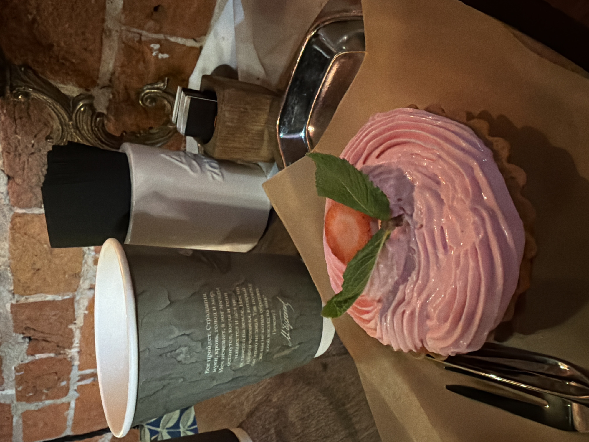 The image captures a scene featuring a dessert and a beverage, likely in a café or restaurant setting. The dessert is a tart with a pink, creamy topping, garnished with a slice of strawberry and mint leaves. It's presented on a piece of brown parchment paper. A coffee cup with a white lid, covered in Russian script, is positioned near the tart. Additional items include a white container filled with black napkins, a wooden napkin holder with some napkins, and a small silver tray. A brick wall can be seen in the background. Based on the Russian text on the coffee cup, it is likely the city is Moscow, Russia.