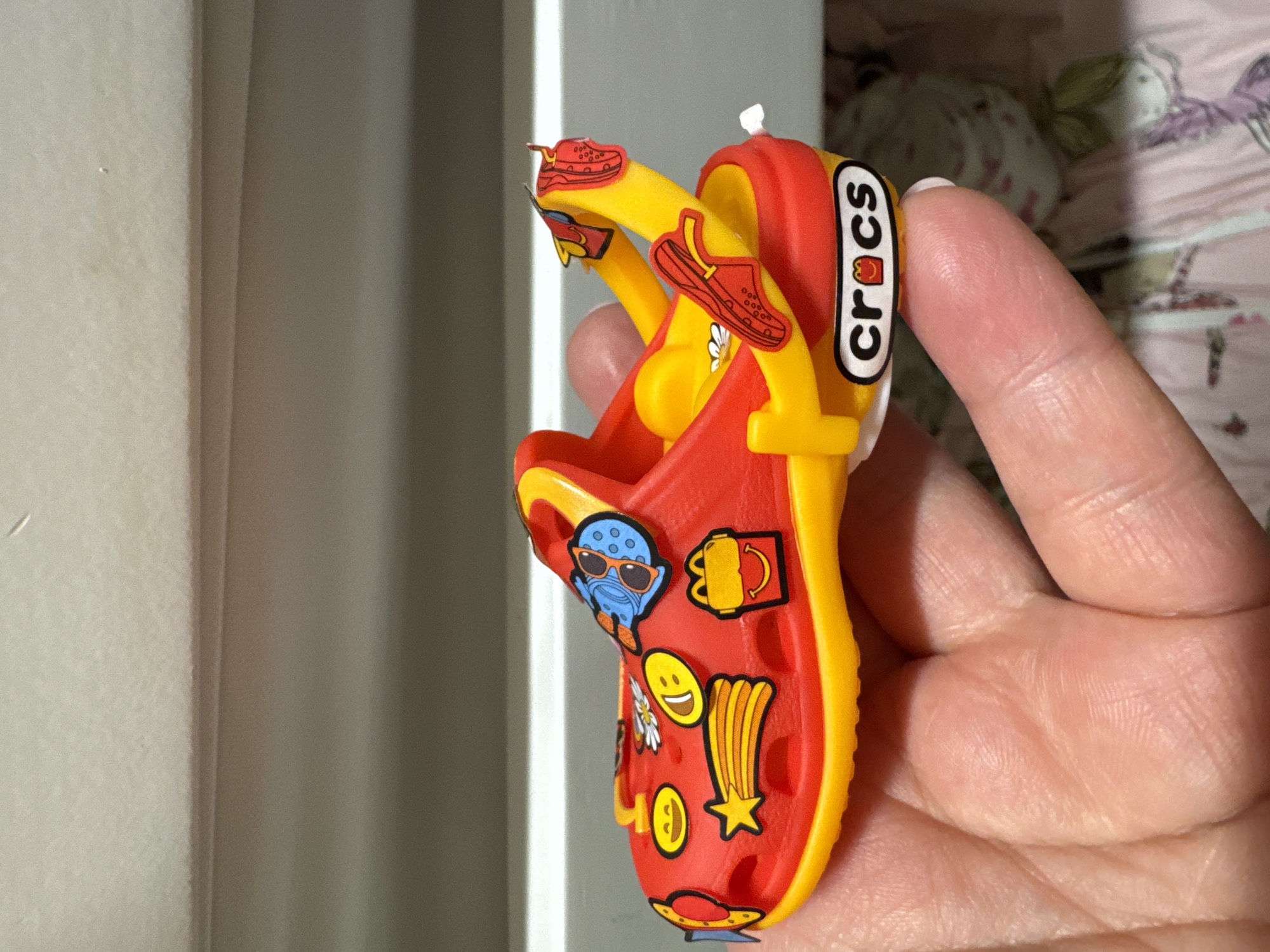 This is a close-up, indoor, eye-level shot of a person's hand holding a red and yellow children's Croc shoe adorned with various colorful Jibbitz. The Jibbitz include cartoon characters, smiley faces, a shooting star, and what appears to be a McDonald's Happy Meal box. The Croc shoe has the "Crocs" logo on the side with a Happy Meal character replacing the "o." The background is softly blurred, showing what appears to be furniture or a wall. There is no indication of the city of Clonmel, Ireland, within the image.