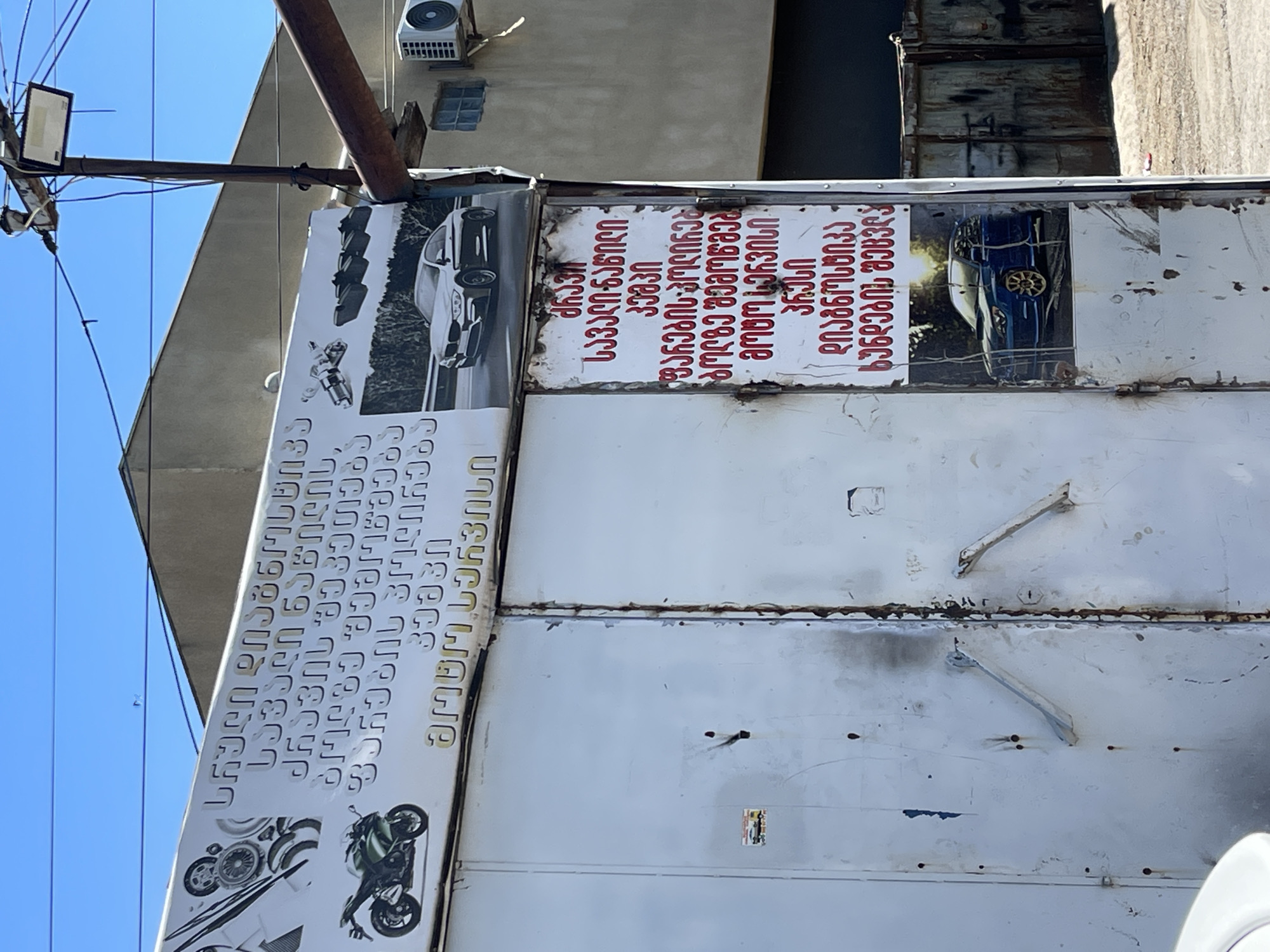 The image shows a sign in the city of Tbilisi, Georgia, attached to a large metal door or gate. The sign features Georgian text and images related to car services, including photos of cars and motorcycles. The text advertises services such as car repair, diagnostics, and wheel alignment. The overall appearance of the scene suggests an automotive shop or service center.