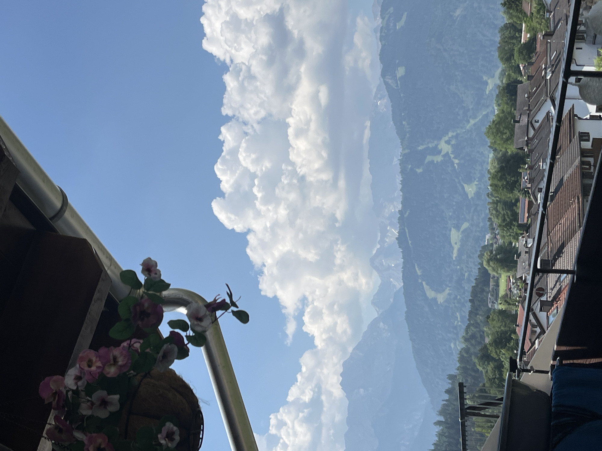 The image shows a view from a balcony in Garmisch-Partenkirchen, Germany, looking out towards a mountainous landscape. In the foreground, a hanging basket of pink and white flowers is visible, partially obscuring a metal downspout. Below the balcony, a cluster of houses with terracotta roofs can be seen. Beyond the houses, rolling green hills covered in trees lead up to imposing, hazy blue mountains. The sky is a bright blue with scattered white, fluffy clouds. A ski slope is faintly visible on one of the mountain sides. The overall scene is one of a serene, scenic vista with a residential foreground. There are no people visible in the image.