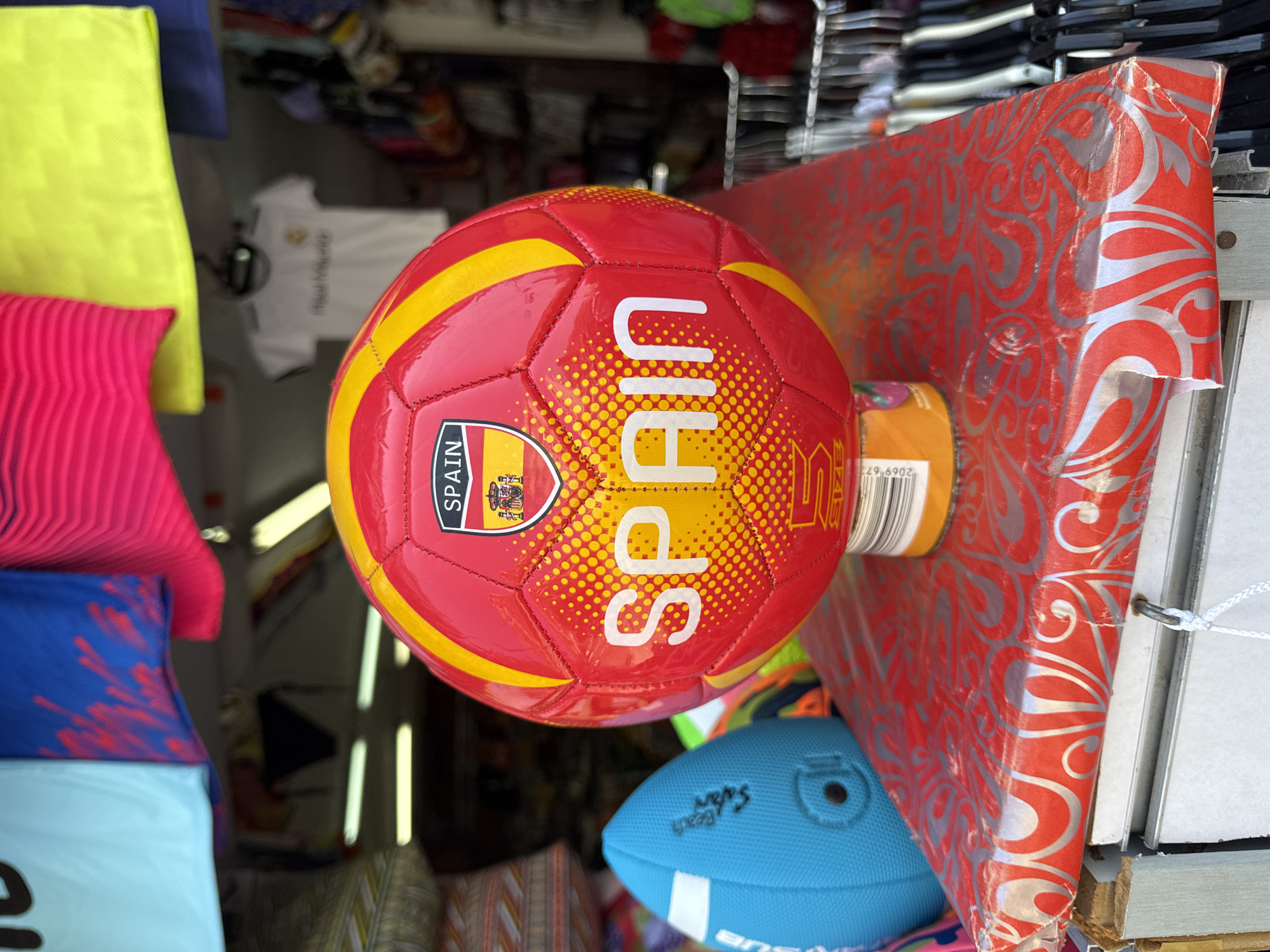 In Blanes, Spain, a red and yellow soccer ball with the word "SPAIN" emblazoned on it sits on a red and silver patterned surface. Behind the ball, various colorful shirts and athletic wear hang, suggesting a retail or market environment. To the left, a blue object, possibly a smaller ball or equipment, is visible. The overall scene appears to be an outdoor stall or shop displaying merchandise.