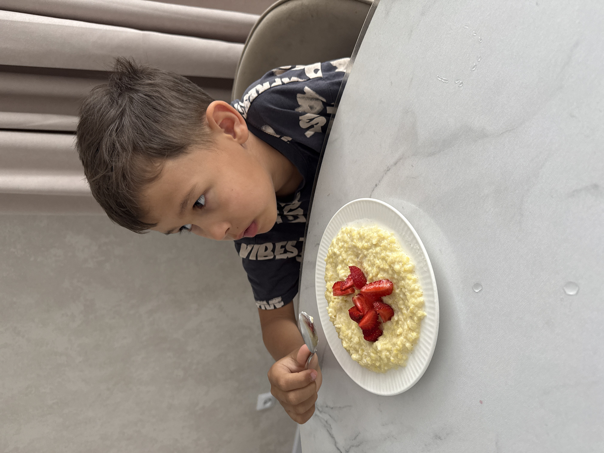A young boy sits at a marble table in an interior space. He is looking to his right with a bowl of oatmeal topped with sliced strawberries in front of him. He is holding a spoon in his right hand, poised as if to take a bite. His dark hair is short and tousled. He is wearing a dark t-shirt with a graphic print. The background shows a textured wall and draped curtains. The image does not provide enough information to determine its location in Nizhnyarkeyevo, Russia.