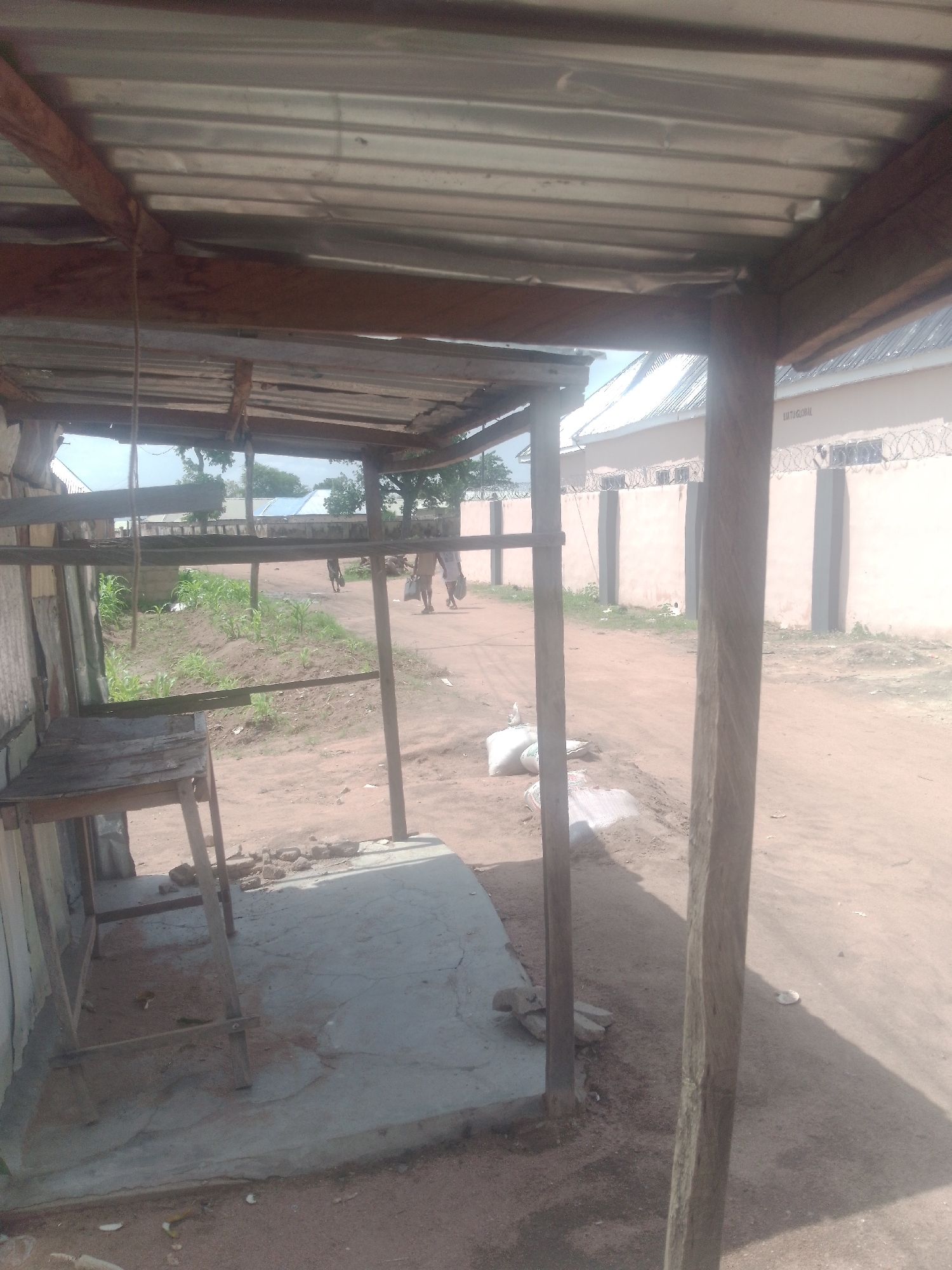 The image depicts an outdoor scene in Jauro Yinu, Nigeria. In the foreground, a makeshift shelter with a corrugated metal roof supported by wooden beams and posts is visible. Underneath the shelter, a simple wooden table or stand rests on a concrete slab. To the left of the shelter, part of a wall is visible.
The background shows a dirt road and a parcel of land with young green crops. Further back, there are buildings with corrugated metal roofs and a tall wall topped with barbed wire. Several people are walking on the road in the mid-ground, some carrying bags. There are also two cows visible further down the road. The overall environment appears to be rural or semi-urban, with elements of construction and agriculture present.