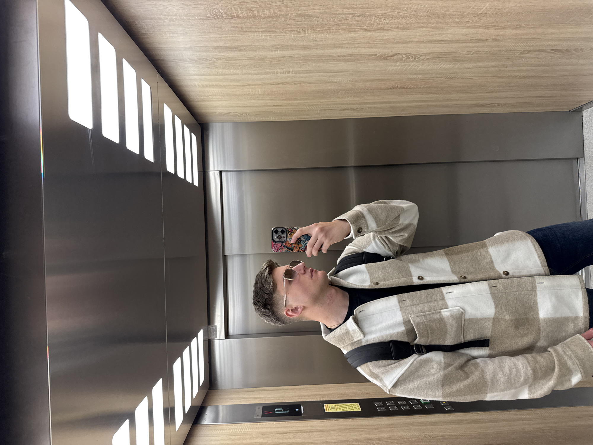 A man is taking a selfie in an elevator. The elevator has stainless steel walls and ceiling with a wood panel above. The elevator has a row of square recessed lights running down one wall. The man is wearing sunglasses and a plaid jacket with a black undershirt. He has a backpack. The location is unknown and there is nothing to indicate Plovdiv, Bulgaria.
