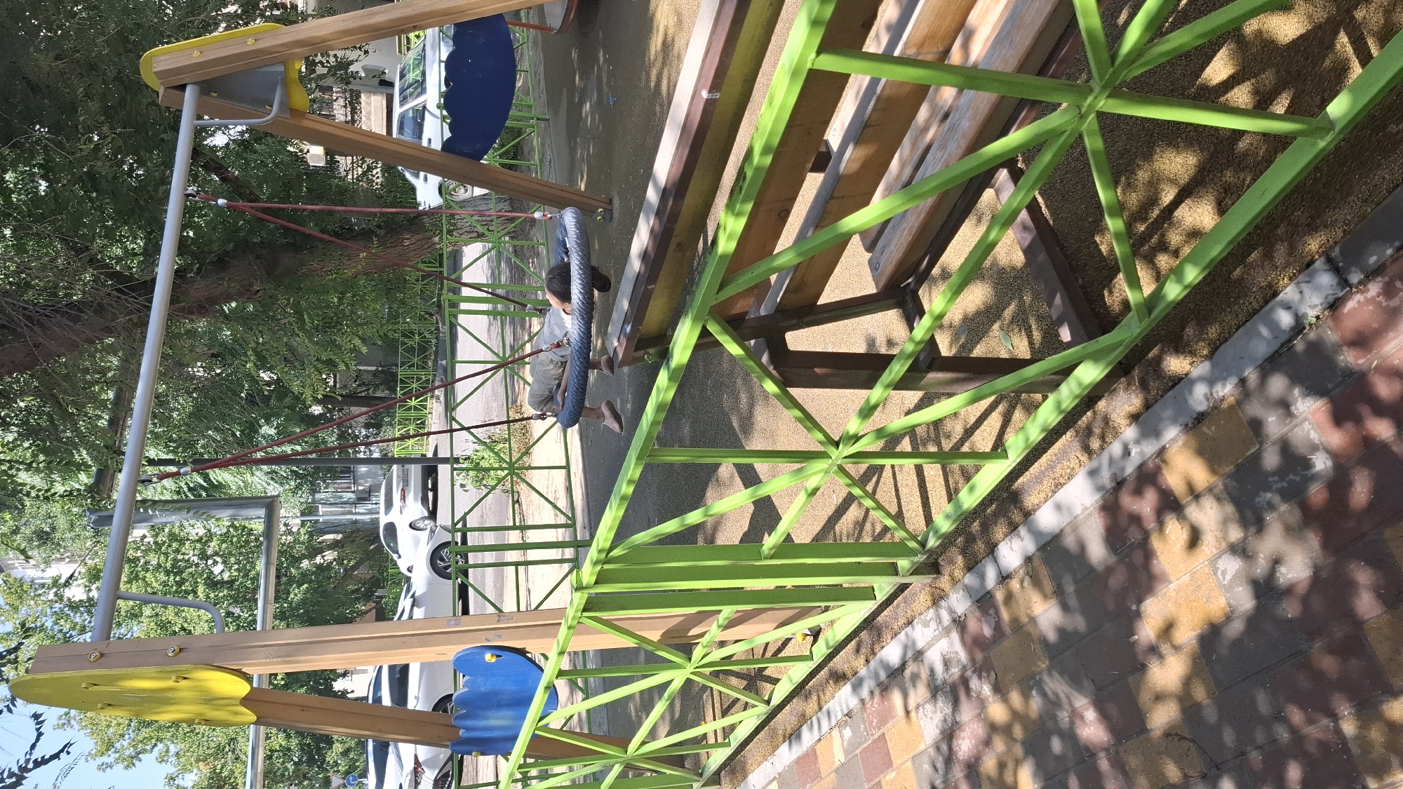 A child is lying on a tire swing at a playground in Shymkent, Kazakhstan. The playground features a wooden swing set with yellow accents and a blue decorative piece. The swing set is surrounded by a green metal fence. In the background, trees and buildings are visible, along with several parked cars on a street. The ground of the playground is covered with soft, brown material, and a paved walkway with a patterned brick design is in the foreground. The scene is illuminated by sunlight, casting shadows from the trees and fence.