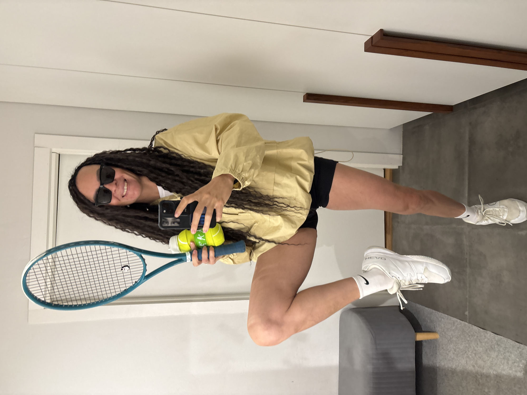 A young individual with long, dark dreadlocks and sunglasses is captured in a full-body mirror selfie, smiling brightly. They are wearing a light yellow anorak-style jacket over a white top, black shorts, white athletic socks, and white sneakers. The person holds a black smartphone in one hand to take the photo, while the other hand grasps the handle of a teal and white tennis racket. Tucked into the racket strings are two yellow tennis balls, one of which visibly features a "KAPPA" brand logo. One of the white sneakers has "HEAD" emblazoned on it, and a Nike swoosh logo is visible on a sock.
The indoor setting features light-colored walls, including a white door frame on the left, and a grey, possibly concrete, floor. Two dark wooden horizontal beams or decorative elements are fixed to the wall or ceiling. In the lower right corner, a grey upholstered bench or ottoman with a wooden support is partially visible. The overall mood is cheerful and active, suggesting a sporty readiness. The location is Chaikovsky, Russia.