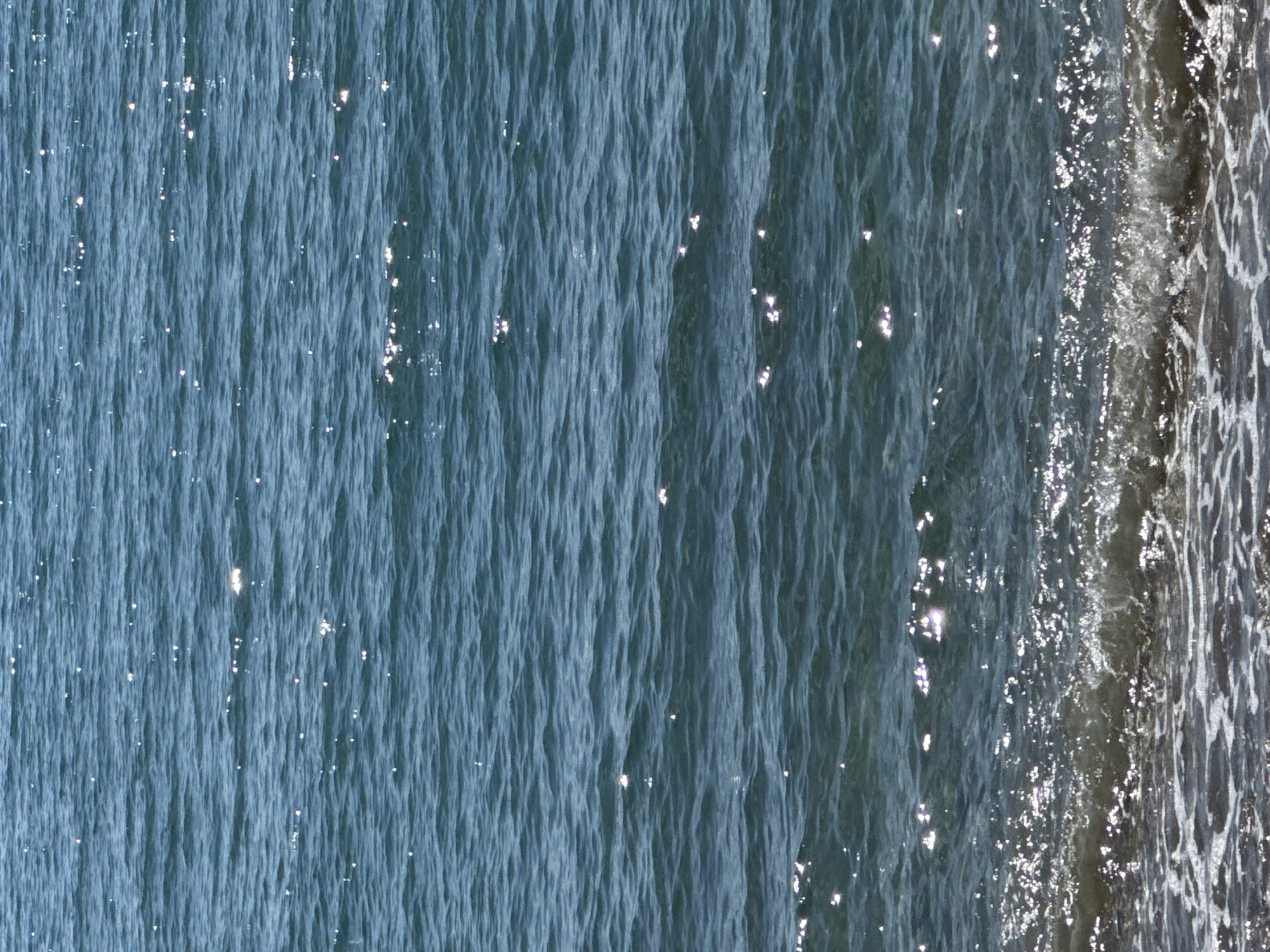 The image shows a body of water, likely the Adriatic Sea, with the shoreline visible on the right side. The water is a blue-grey hue and slightly agitated, as evidenced by the ripples and reflections. The shoreline consists of foamy water with an underlying substrate that appears grey or dark. The location is likely in the city of Bar, Montenegro, given the prompt's location specification. No people, other subjects, or interactions are visible in the image.
