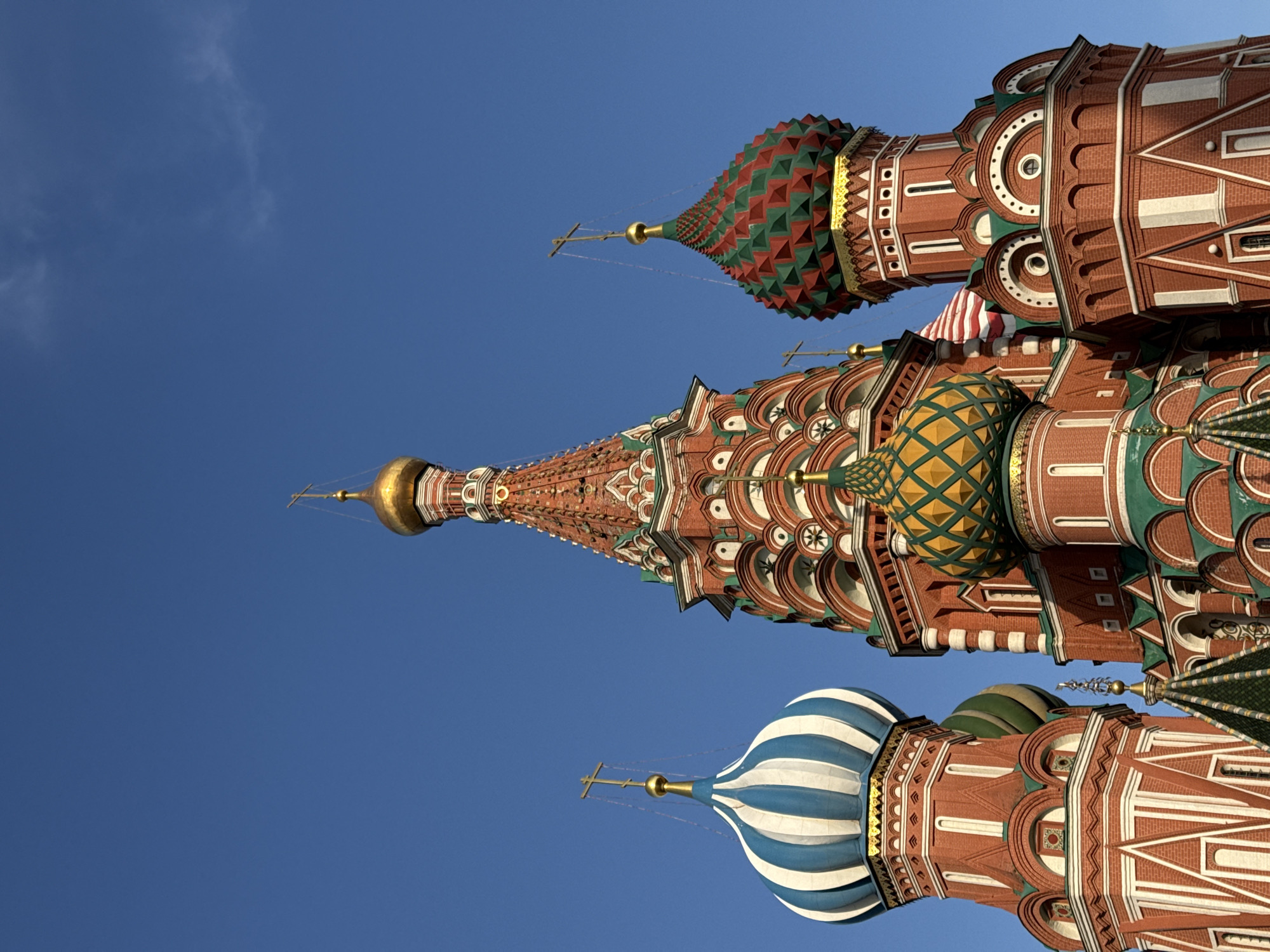 The image shows a low-angle view of St. Basil's Cathedral in Moscow, Russia. The cathedral's colorful onion domes are prominently featured against a clear blue sky. The domes are decorated with various patterns and colors, including red, green, blue, and gold. The architectural details of the building's facade are visible, including brickwork, arches, and decorative elements. No people are visible in the frame. The overall impression is one of a clear, sunny day showcasing the iconic architecture of St. Basil's Cathedral.