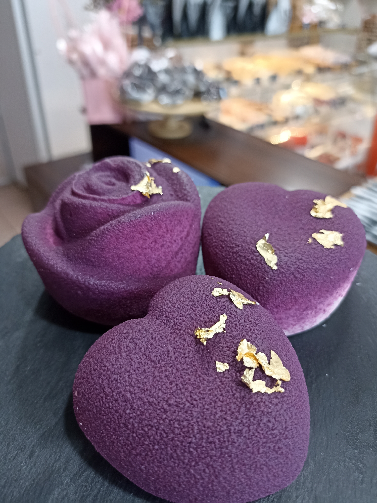The image shows three purple-colored, textured desserts, likely pastries, arranged on a dark gray or black surface. One dessert is shaped like a rose, while the other two are shaped like hearts. Each dessert is decorated with small pieces of gold leaf. The background is blurred, but it appears to be a bakery or pastry shop, with display cases filled with various baked goods visible in the distance. The location of the city is Unknown, Unknown.
