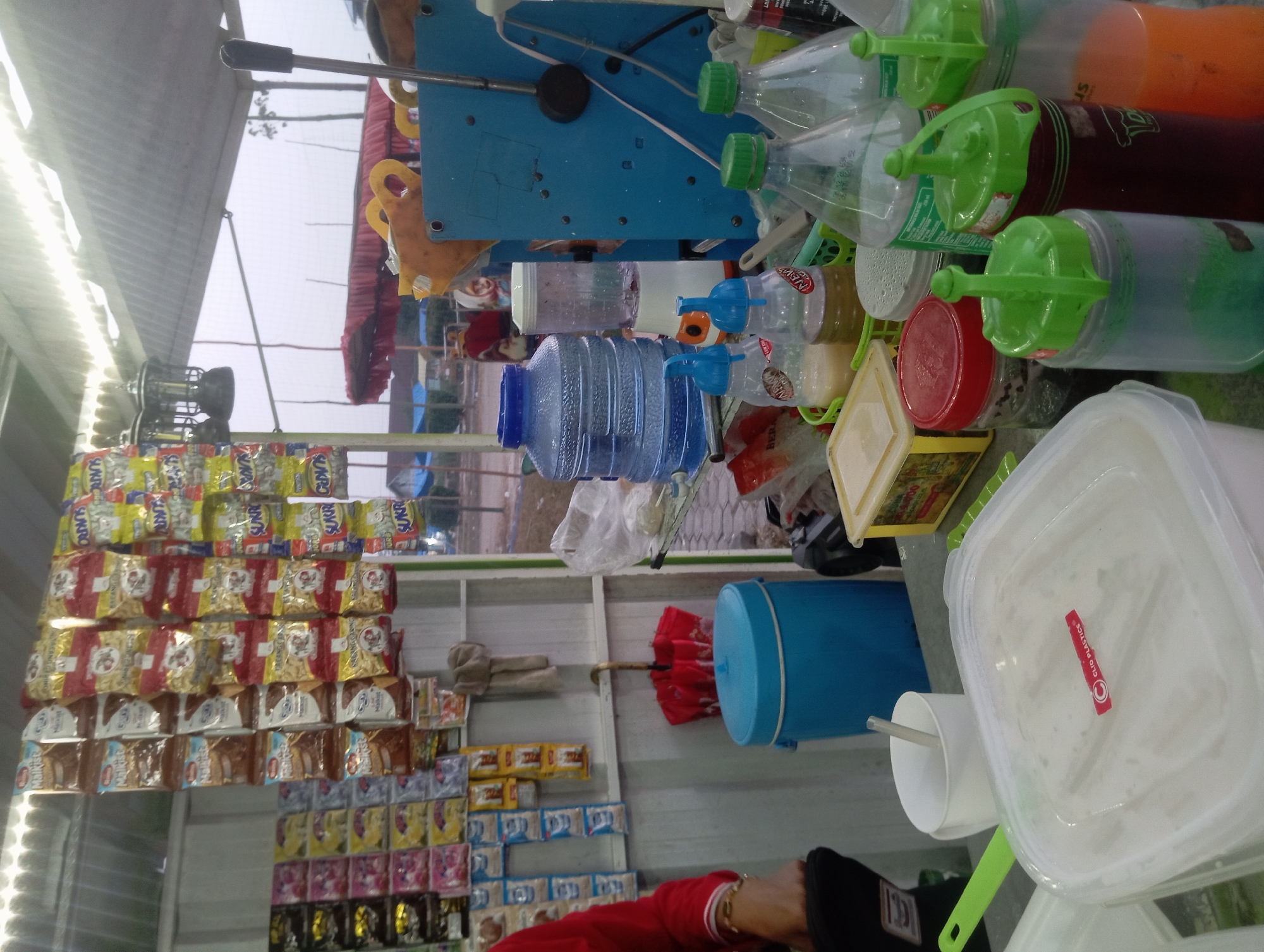 The image depicts an outdoor stall, likely a roadside kiosk, in Way Dente, Indonesia. The stall is organized, with various items displayed for sale. Suspended from the corrugated metal roof are numerous individually packaged snacks, including bags with red and yellow packaging and bags with text that appears to be in Indonesian. Below the snacks, a blue plastic container with a red umbrella folded on top is visible, along with a white mug and a green ladle.
In the foreground, a large clear plastic container filled with a white substance sits on a table. Adjacent to it are several bottles with colorful liquids and green lids, suggesting beverages or condiments. Other items on the table include a yellow container with a lid, and two smaller bottles with blue caps. A person's arm, wearing a red long-sleeved shirt, is visible on the left side of the frame, suggesting they are either the vendor or a customer. The background shows a blurred view of the outdoors, with some structures and vegetation. The overall scene suggests a small, bustling business operating in a public area.