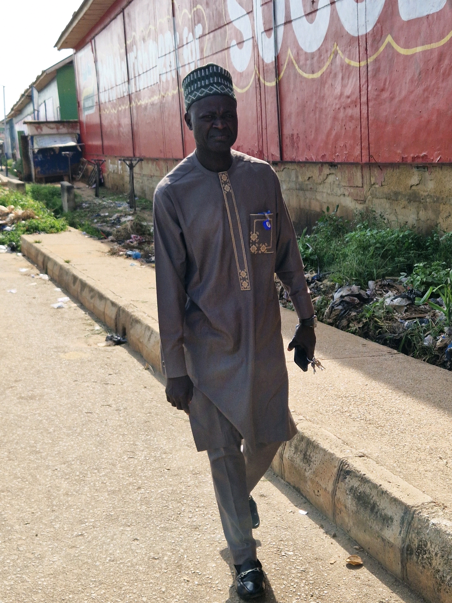A Black man in his 50s, wearing a dark grey traditional outfit with gold trim and a green and black embroidered cap, walks on a paved road. He is holding a set of keys in his right hand, which is gloved in black. The man is looking directly at the camera. Behind him, a red building with large white circular designs and yellow wavy lines painted on it stretches along the side of the road. The area to the right of the road is filled with overgrown green vegetation and scattered piles of trash, suggesting an urban environment. To the left, a sidewalk runs alongside the road, also with some debris visible. The lighting indicates it is daytime, with shadows cast on the ground, suggesting a sunny day. The scene appears to be in an outdoor, possibly commercial or residential, area in the city of Kabong, Nigeria.