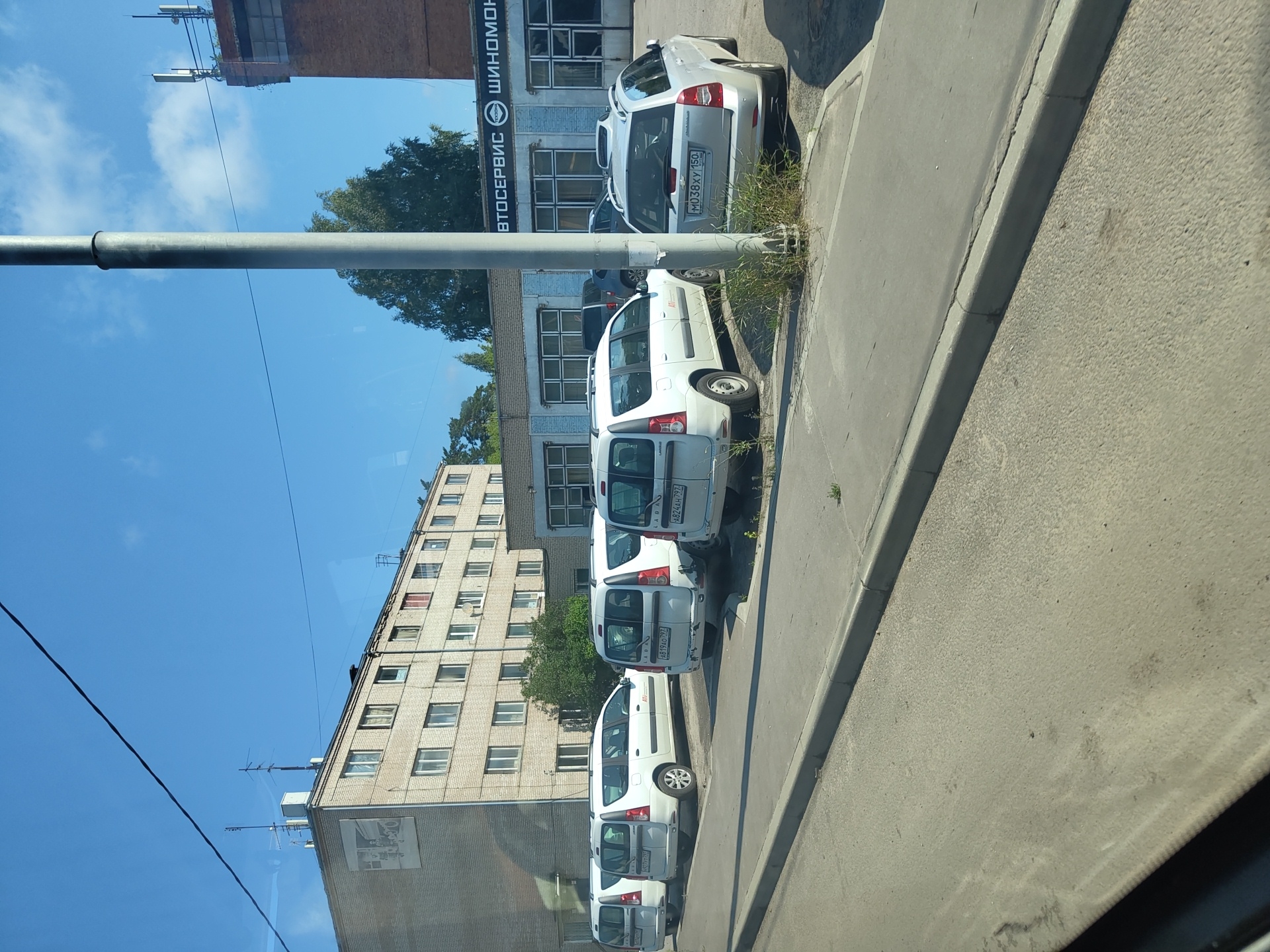 The image depicts a street scene in the city of Zhukovsky, Russia, on a clear day. A light gray, multi-story apartment building stands on the left side of the frame, its brick facade and numerous windows visible. To the right of the apartment building, a low-rise commercial building with a blue and gray facade is visible, featuring several large windows and signage that appears to read "АВТОСЕРВИС" and "ШИНОМОНТАЖ", indicating an auto service and tire fitting shop. A tall, gray utility pole stands prominently in the foreground. Parked along the sidewalk in front of the commercial building are several white Lada Granta station wagons. One silver Chevrolet Aveo is also parked in the vicinity. The street and sidewalk are paved, with some patches of grass growing through cracks. The sky above is a clear, vibrant blue.
