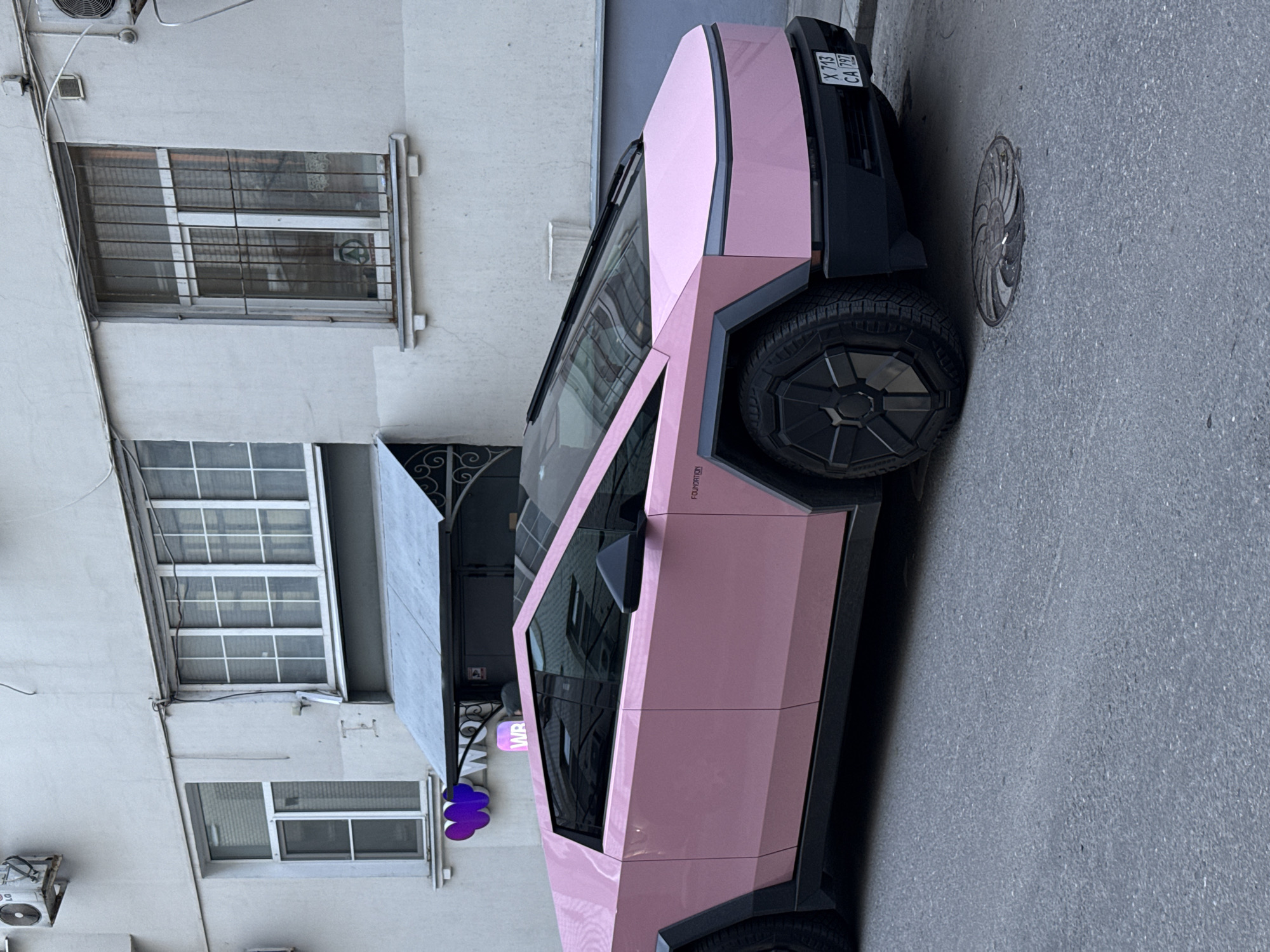 A distinctive pink Tesla Cybertruck is parked on an urban street next to a light-colored building during daytime, under soft, diffused lighting. The Cybertruck's angular body is custom wrapped in a matte pink finish, contrasting with its black wheel arches and multi-spoke black wheels. The word "FOUNDATION" is partially visible in black text on the lower side of the front door. The vehicle's license plate on the front reads "X 713 CA 797", indicating registration in Moscow, Russia.
The building alongside the car features an off-white, textured facade with multiple windows. Some of these windows, particularly on the lower levels, are secured with horizontal metal bars. A faint circular green logo is reflected in the glass behind the security bars of one upper window. An external air conditioning unit is mounted high on the building wall. A small purple sign displaying the letters "WR" and an abstract purple shape is affixed to the building below one of the windows, suggesting a business. The street is paved with asphalt, and a circular, patterned manhole cover is visible near the front of the Cybertruck. No people or other activities are depicted in the scene.