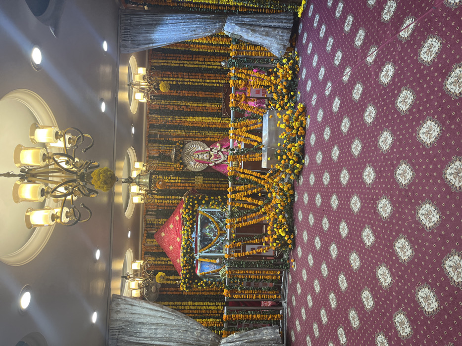 The image depicts the interior of a temple decorated with garlands of marigold flowers. The temple is located at 30.723° N, 76.862° E in Panchkula, India. The scene is dominated by a red carpet with white patterned designs covering the floor. There is a deity statue sitting in a decorated shrine in the back. The walls are adorned with garlands of marigold flowers and there is a large chandelier hanging from the ceiling. Two large curtains, one on each side, are hanging in the background. The shrine has a red canopy and is decorated with white flowers. There is a metallic railing in front of the shrine. There are no people present in the image. The environment is dimly lit with soft, warm lighting.