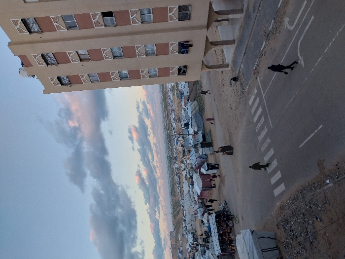 A tall beige building stands beside a vast landscape of tents. Below, a road stretches out, marked with a zebra crossing and directional arrows, where several people are walking or crossing. The sky above is a mix of blue and clouds. The perspective looks down from above, possibly from another building or elevated position.