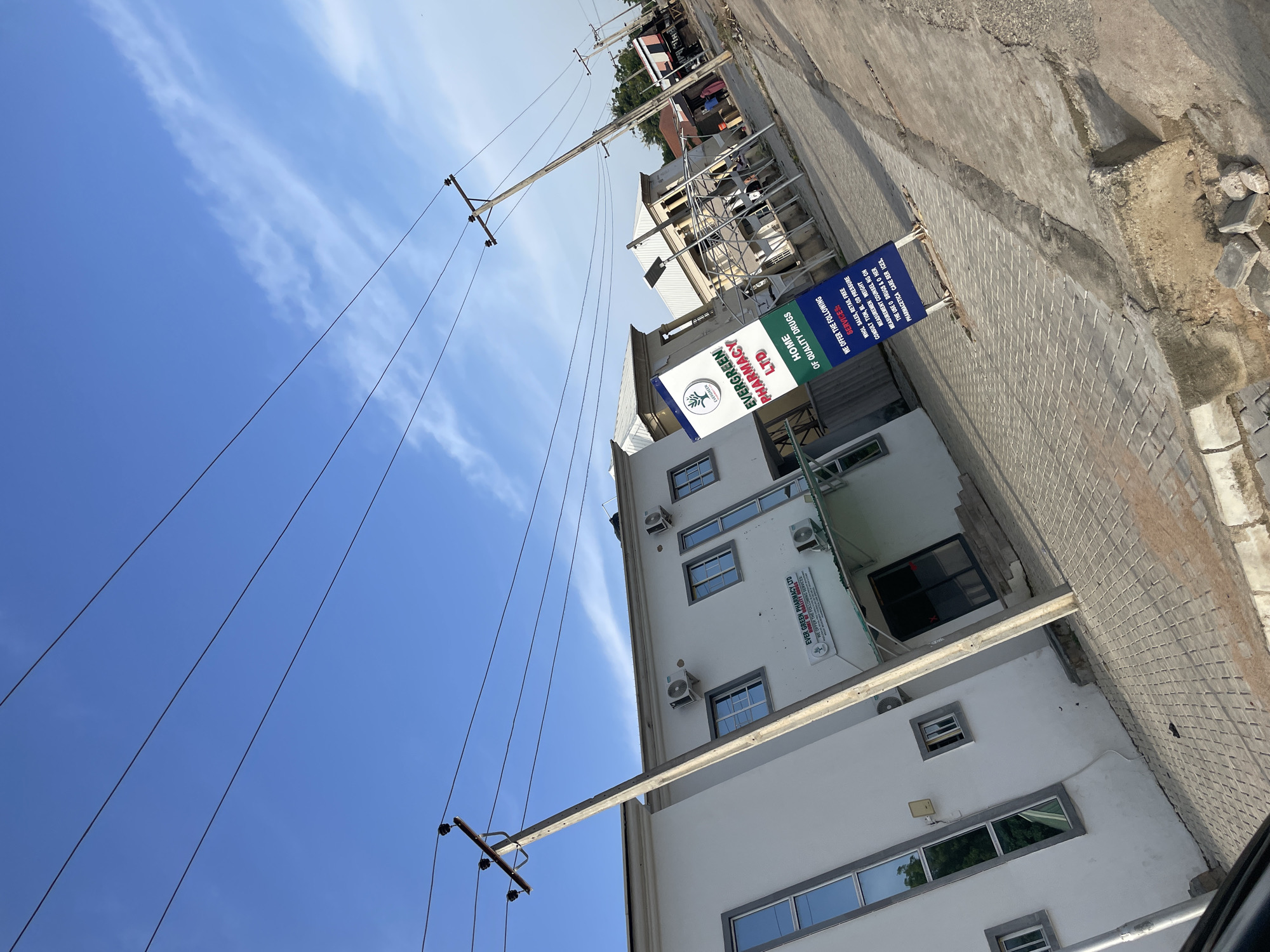 The image shows a street scene in Jalingo, Nigeria, featuring a white building with blue-tinted windows, identified by a sign as "EVERGREEN PHARMACY LTD." The sign also indicates "HOME OF QUALITY DRUGS" and lists services offered. A concrete utility pole with electrical wires runs diagonally across the scene, in front of the building. The ground is paved with interlocking bricks. In the background, other buildings and a few people can be seen, suggesting an urban or semi-urban environment. The sky is blue with scattered clouds, and the lighting indicates it is daytime.