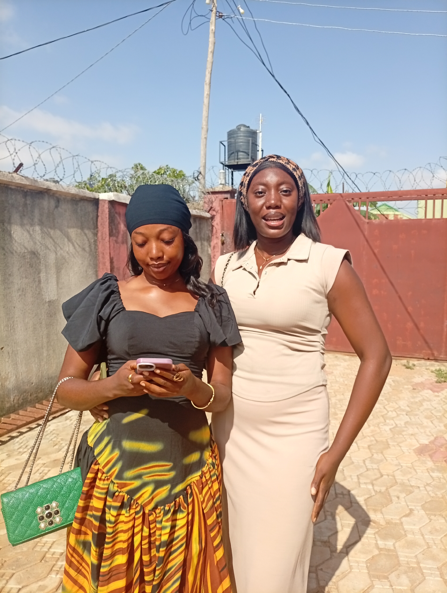 Two women stand close together outdoors in Rido, Nigeria. One woman wears a black head covering, a black top with puff sleeves, and a skirt with yellow, brown, and black patterns. She holds a pink smartphone and wears jewelry on her wrists and fingers. A green quilted purse hangs from her shoulder. The second woman wears a printed headband and a beige collared dress. Behind them is a concrete wall topped with barbed wire, a tall wooden utility pole with wires, and a red gate. A black water tank sits above the wall. The ground is paved with hexagonal tiles. The sky is blue.