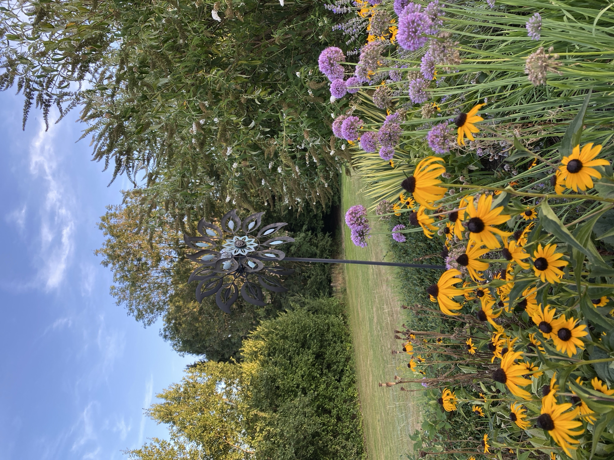 The image depicts a garden with a variety of flowers in bloom under a clear blue sky with scattered white clouds. In the foreground, there is a dense cluster of yellow Black-Eyed Susans and purple Alliums. Behind them, a tall, dark metal wind spinner shaped like a flower stands on a pole. In the background, a lush green lawn stretches towards a dense line of trees. The overall impression is of a well-maintained garden scene. The image does not provide any information about the city of Helmenzen, Germany, or any people.