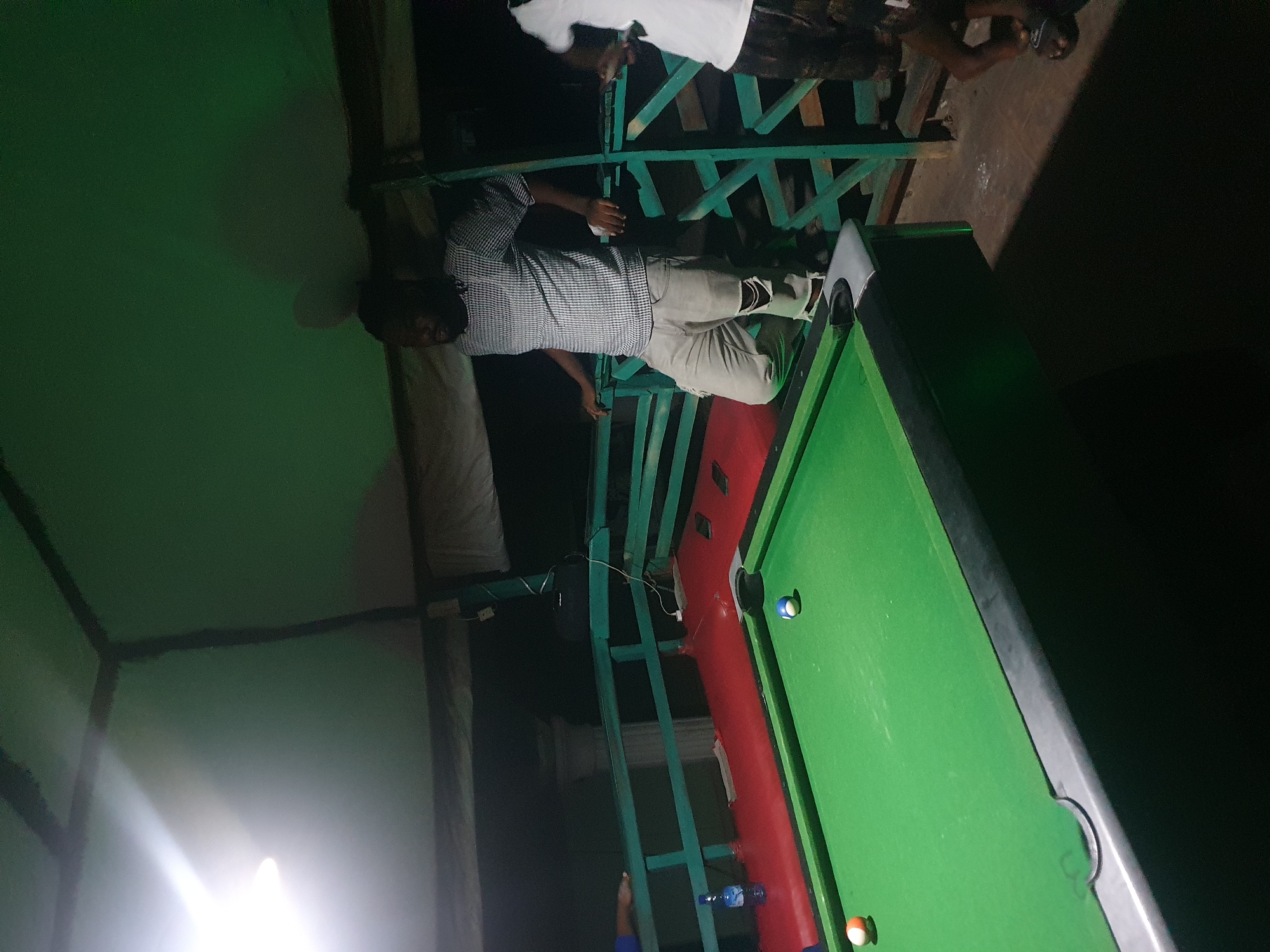 In Lokoja, Nigeria, at night, a billiard table is set up outdoors under a green canopy. A man in a checkered shirt and ripped gray pants leans on a turquoise railing near the table. Another person, wearing a white shirt and patterned shorts, stands to the right, partially visible. Two billiard balls, one orange and white and another blue and white, are on the green felt of the table. A blue bottle of water sits on a red surface beside the railing, near a person's hand reaching over the railing. The scene is dimly lit, with a bright light source visible in the upper left corner.
