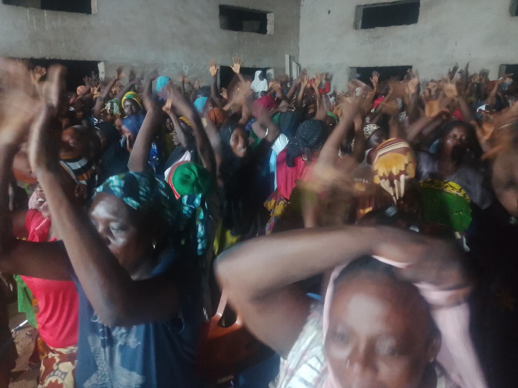In an indoor setting, possibly a community hall or place of worship, a diverse group of people, predominantly women, are captured in a moment of communal engagement. The lighting is dim, suggesting an evening or overcast day. The individuals are in close proximity, many with their arms raised, indicating active participation in a song, prayer, or dance. Their faces show a range of expressions, from joyous exuberance to focused devotion. The attire is varied, with many women wearing brightly colored headscarves and traditional African prints. The background consists of unadorned concrete walls, with what appear to be windows or openings suggesting a simple, functional structure. The overall atmosphere is one of shared energy and collective experience.