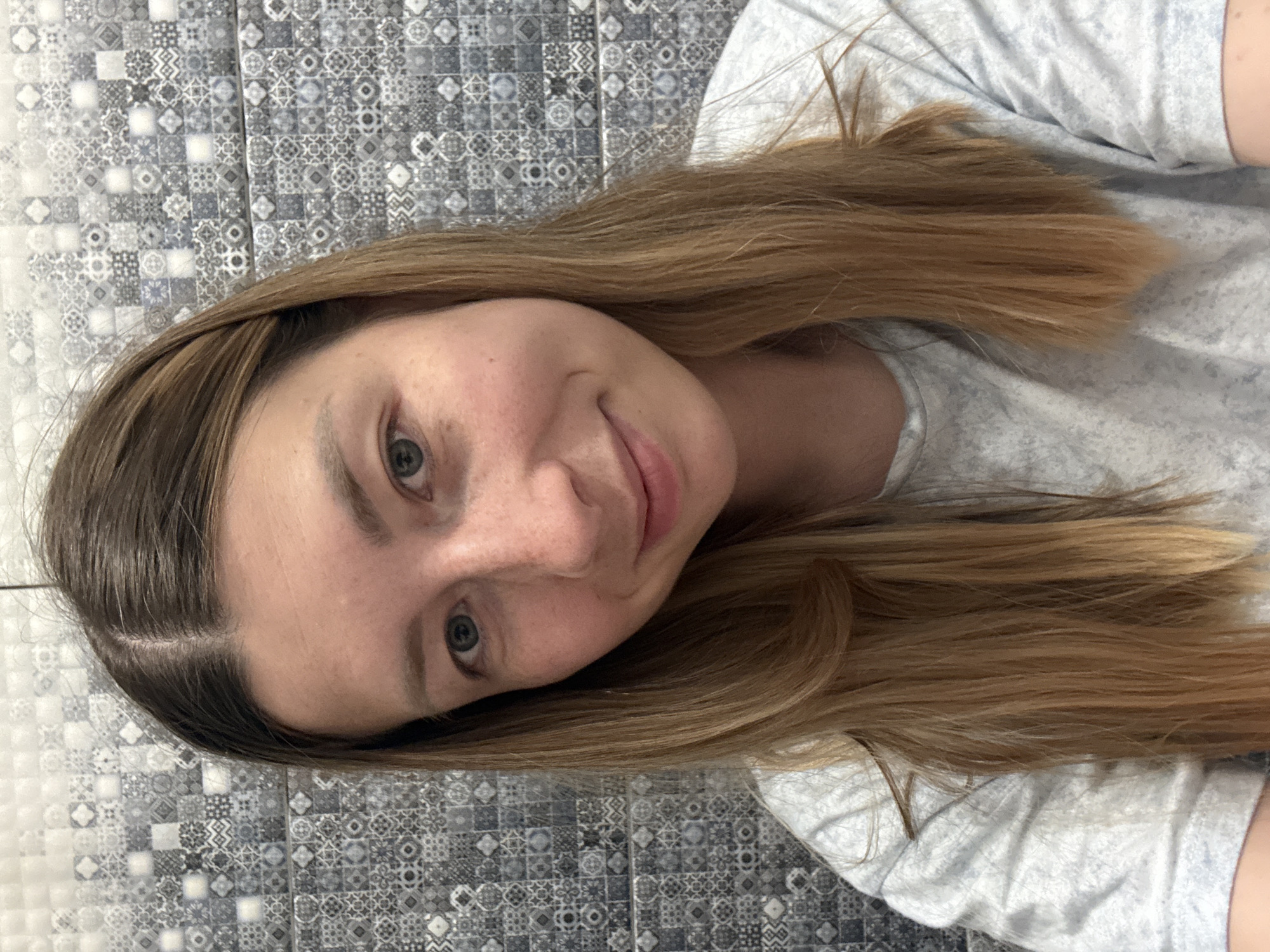 A young woman with long, light brown hair is positioned against a tiled background. She is wearing a light-colored shirt. The tiles behind her are patterned in a gray and white mosaic design. Her expression is neutral with a slight smile. The image is rotated, giving a skewed perspective of the scene. There is no indication of location in Moscow, Russia.