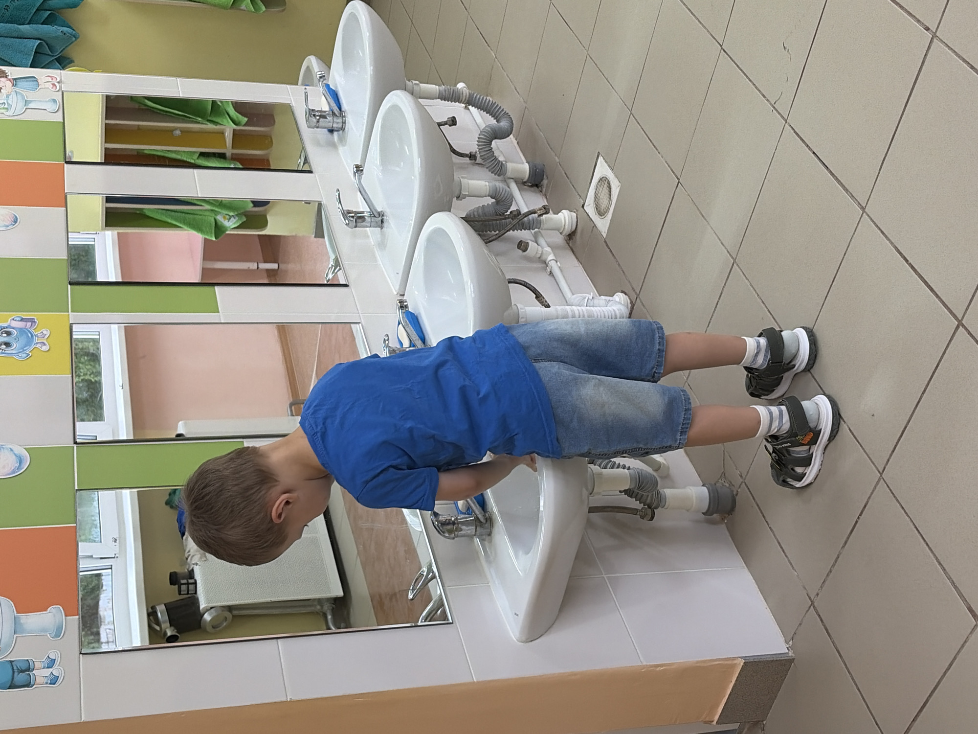 A boy is washing his hands in a white porcelain sink in a tiled bathroom. The boy is wearing a blue t-shirt, jean shorts, and striped socks with black and gray sandals. The bathroom has three sinks in a row, each with a mirror above it. The mirrors are framed by colorful tiles. The floor is tiled in beige. The location is Chaikovsky, Russia.