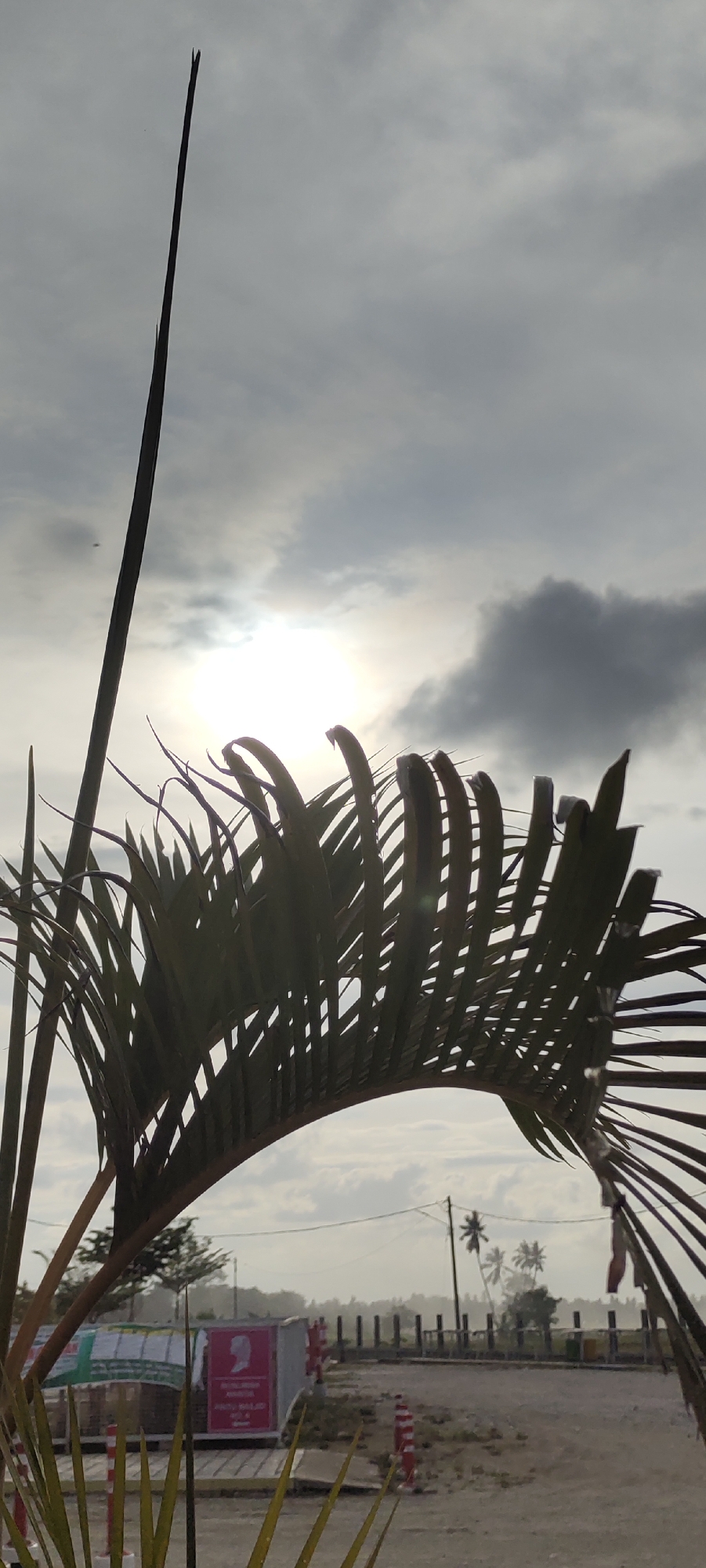 The image, taken in Meunasahaluedrienlhoksukon Barat, Indonesia, features a palm frond in the foreground, silhouetted against an overcast sky. The sun is obscured by clouds, creating a diffused light. Behind the palm, there's a structure or building with signage, and further back, a line of posts or barriers can be seen along a road or pathway, with palm trees in the distance. The overall scene appears somewhat hazy.