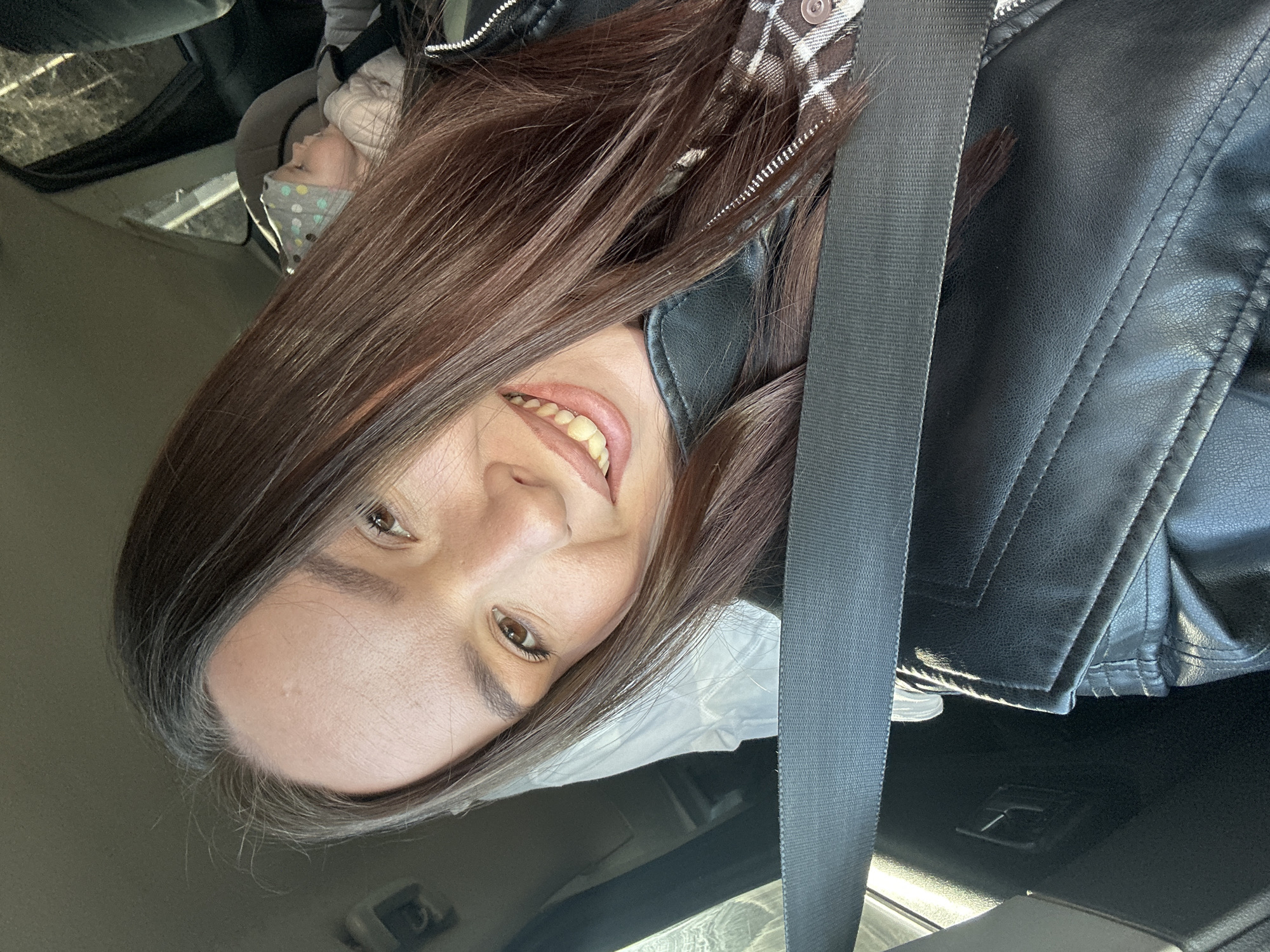 The image, rotated 90 degrees counter-clockwise, captures a woman and a child inside a car, likely in Chaikovsky, Russia. The primary subject is a woman in the foreground, smiling broadly at the camera with her mouth open, revealing her teeth. She has long, dark brown hair and brown eyes. She is dressed in a black faux leather jacket layered over a brown and white plaid shirt, with a black seatbelt extending across her torso.
In the background, to the woman's left, a young child is visible in a car seat. The child is wearing a light-colored face mask with polka dots and appears to be looking upwards or towards the left. The car's interior is dark, with visible upholstery and the black seatbelt as a prominent feature. Natural light suggests the scene takes place during the daytime. The woman's open smile conveys a happy or content emotion. No discernible text is visible within the image.