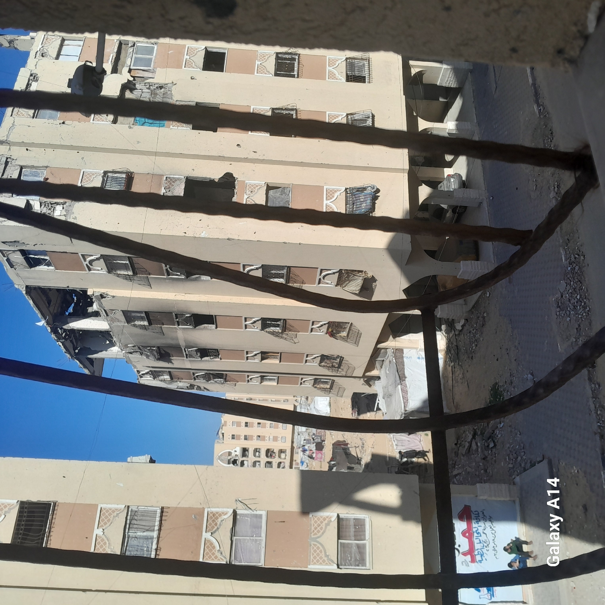 The image shows buildings in Khan Yunis, Palestinian Territory, viewed through a window or balcony railing. The buildings are multi-story, with damage evident on at least one building, including missing facade sections and charred areas. The sky is clear and blue. Below, on the ground level, there is a sign written in Arabic above two figures; and the edge of an area of piled debris is also visible. The lettering "Galaxy A14" is visible in the bottom right corner of the image.