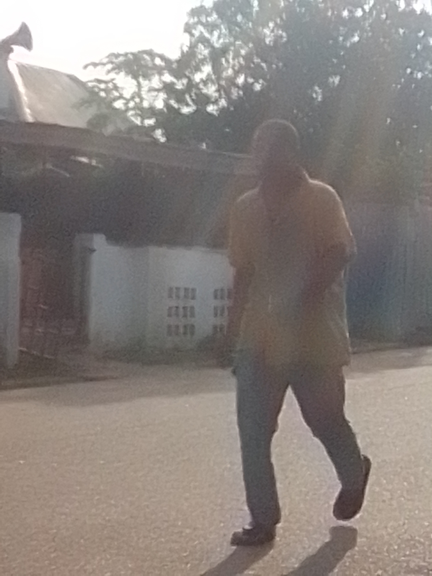 A person, seen from the waist down, walks on a paved road in Liji Hill, Nigeria. The person is wearing dark pants and dark shoes. The background shows a building with a white facade and rows of windows, and a tree-lined area. The image is blurry, suggesting it was taken from a distance or with a low-resolution camera. The lighting suggests it is daytime, possibly late afternoon, given the long shadow cast by the person. There are no other people or vehicles clearly visible in the image. The overall scene depicts an outdoor urban environment.