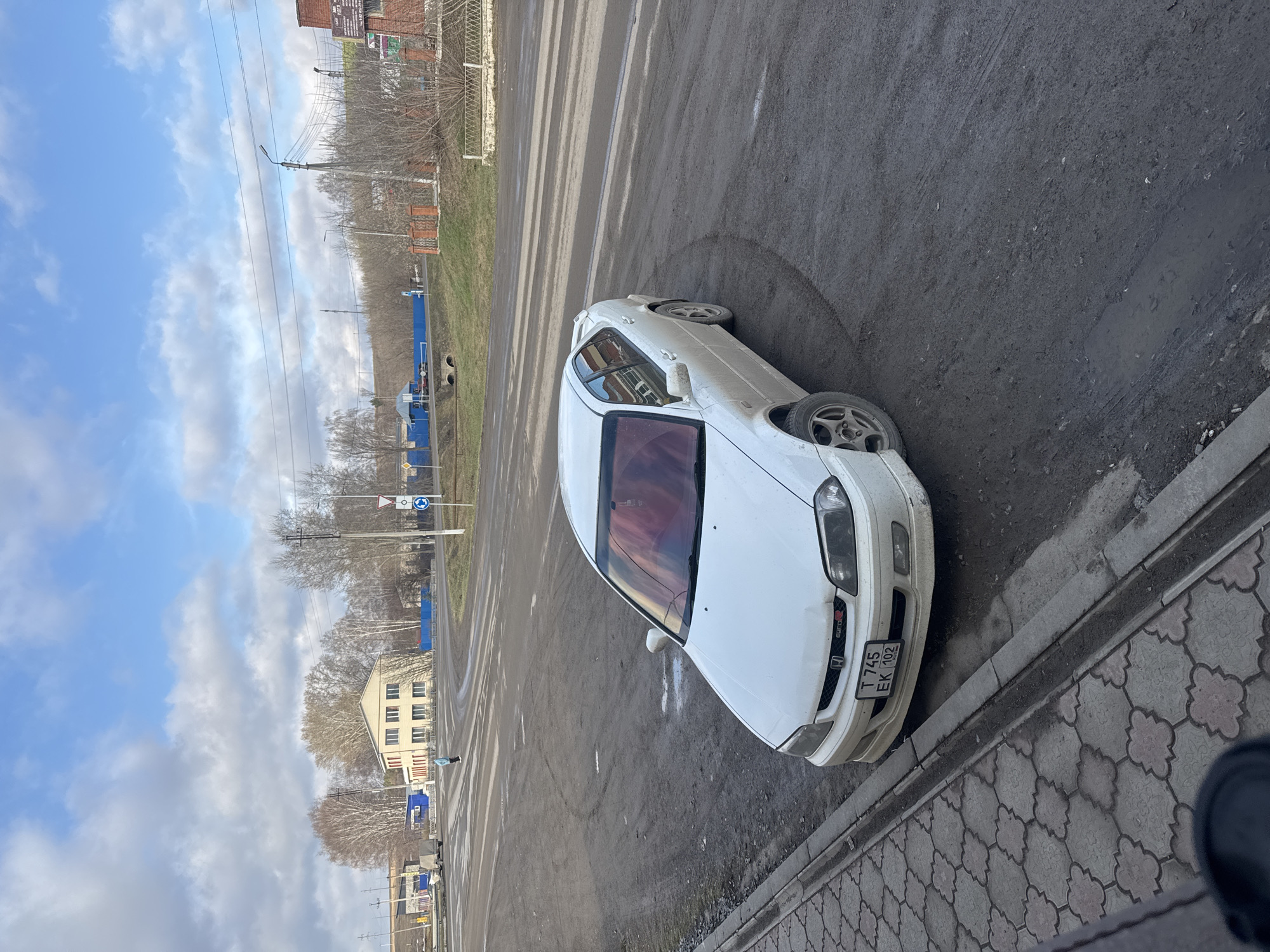 A white Honda Euro R coupe is parked on the side of a street in Mesyagutovo, Russia. The car is situated on a patch of asphalt adjacent to a sidewalk made of patterned grey and white pavers. The street surface shows signs of wear and discoloration. In the background, there is a view of trees without leaves and a building with multiple windows. Blue utility structures are visible along the roadside, and power lines stretch across the sky. The sky is partly cloudy. The license plate is partially obscured but appears to read "T 745 EK 102". There are no people immediately visible near the car. The environment suggests an urban setting in a state of disrepair.