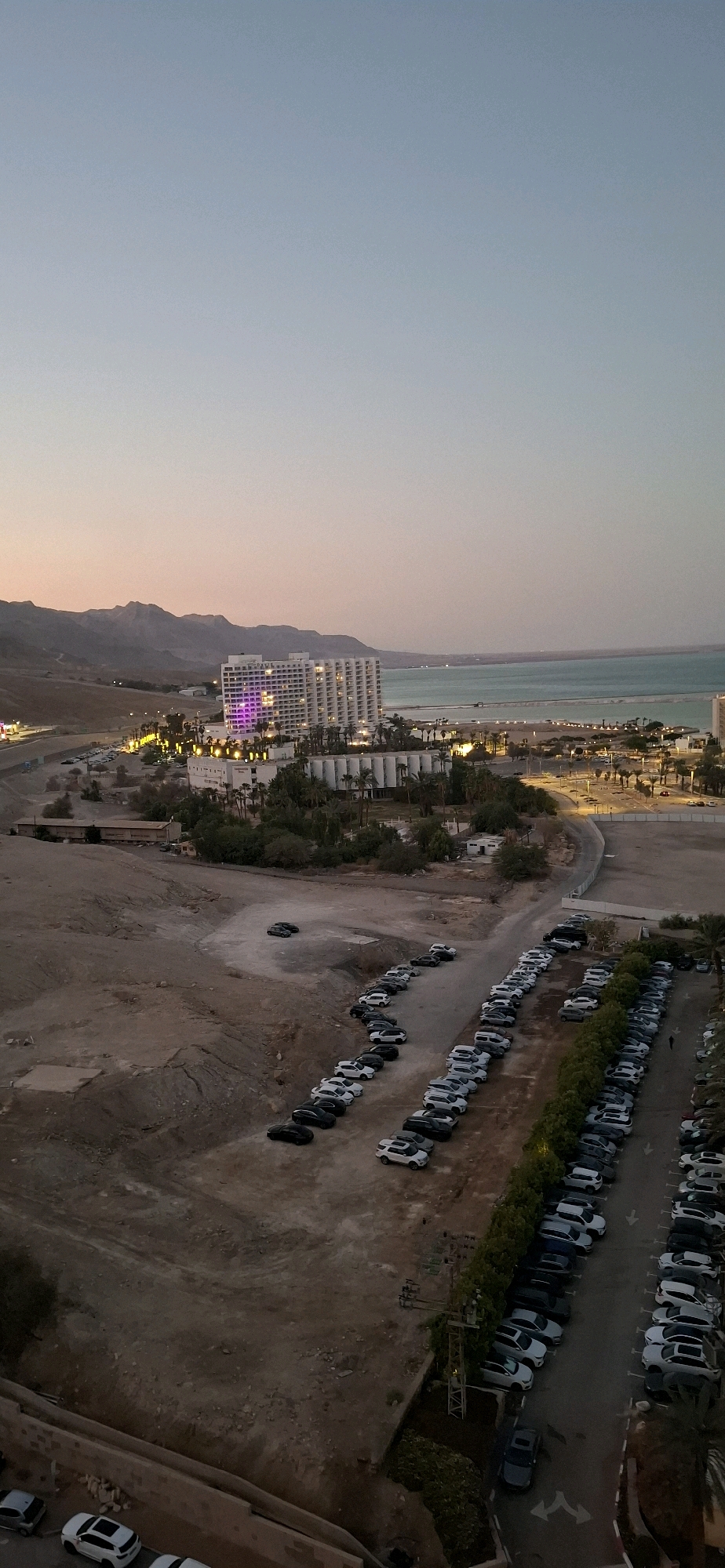 This is an aerial, vertical, full shot of a hotel resort in Tamar Regional Council, Israel, at dusk. The hotel, a multi-story white building with many windows, is illuminated with purple and yellow lights. It is situated on a coastal area with a calm sea in the background. The landscape is arid with brown hills in the distance. Numerous cars are parked in rows in dirt and paved parking lots. Palm trees and other vegetation are scattered throughout the resort area. The sky transitions from a pale blue at the top to a soft orange and pink hue near the horizon, indicating the end of daylight. There are no visible people in the image.