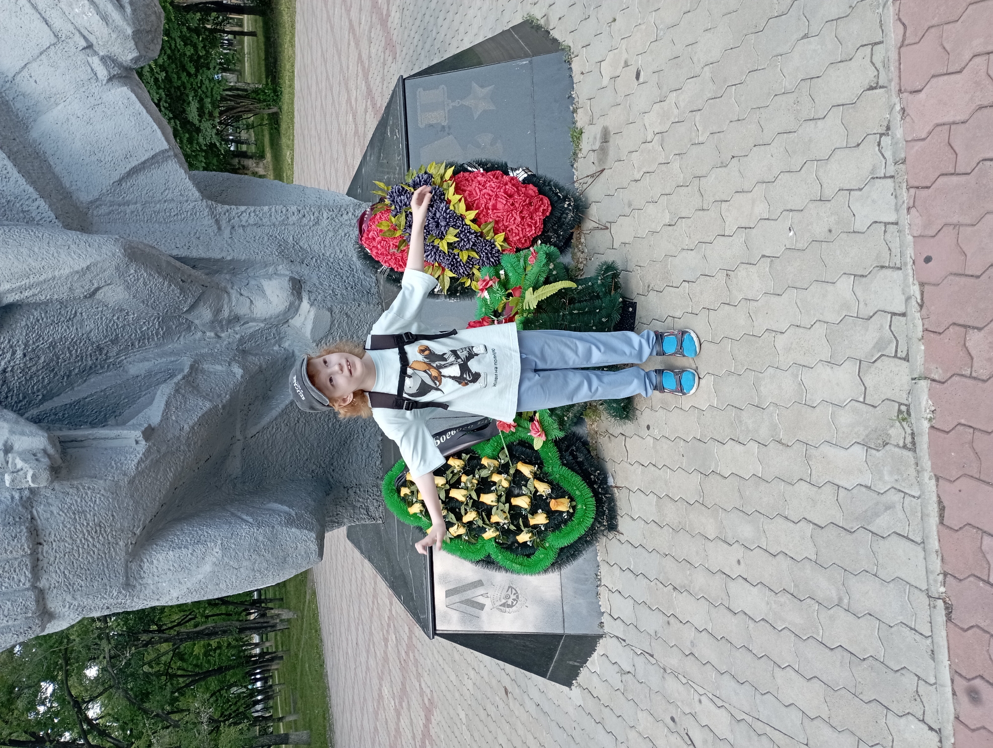 A young child with auburn hair, wearing a light blue t-shirt with a cartoon graphic, light blue pants, a grey baseball cap, and blue sneakers, stands in front of a large, grey, abstract monument. The child has their arms outstretched, palms up, and is looking to their right. Two memorial wreaths are placed at the base of the monument, one to the child's left with yellow flowers and green foliage, and another to the child's right with red and purple flowers and green foliage. The monument and wreaths are situated on a paved walkway made of interlocking grey bricks. In the background, trees and grass are visible, suggesting an outdoor park or public space. The scene appears to be located at the Eternal Flame monument in Blagoveshchensk, Russia.