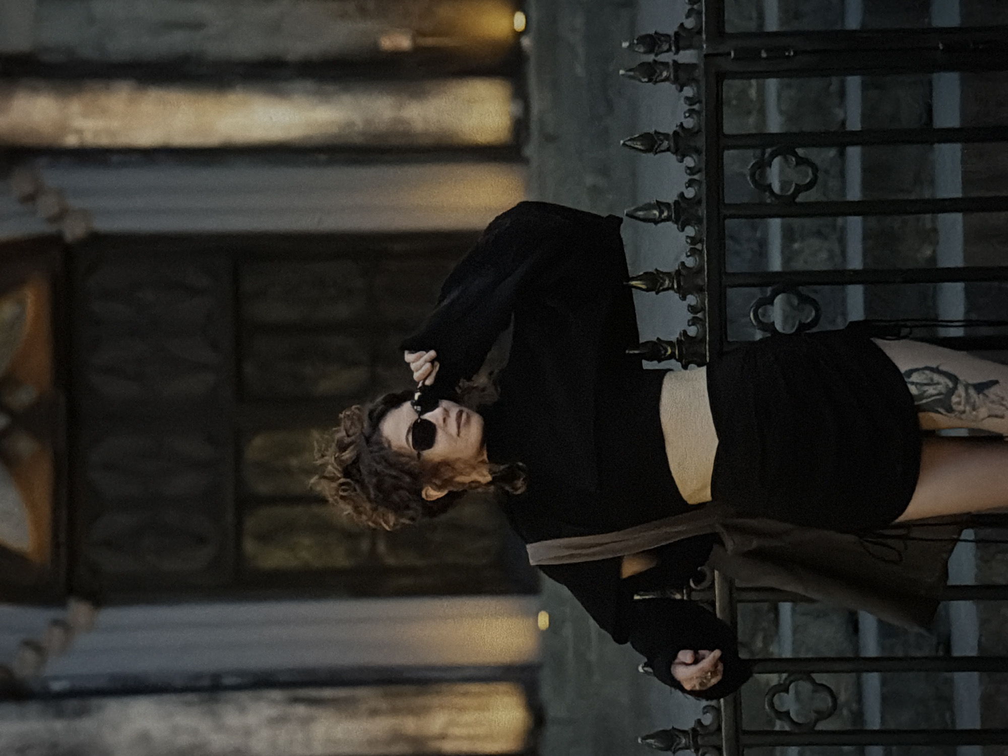 A woman with curly brown hair and sunglasses, wearing a black crop top and mini skirt, stands in front of a metal gate. Her left leg is visible and has tattoos. She has one hand near her face, with the other resting on the gate. Behind her is a stone building with a large arched doorway, bathed in warm light. The setting appears to be outdoors, likely in an urban environment. The location in the city of Đà Nẵng, Vietnam cannot be determined.