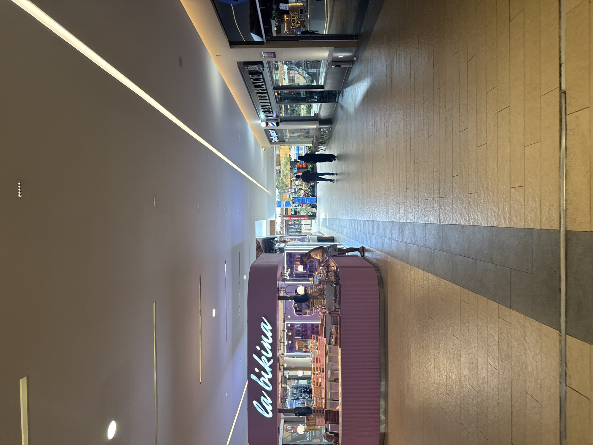 The image is an interior shot of a shopping mall in Puebla, Mexico. The scene is a long corridor with stores on either side and people walking through it.
On the left side of the corridor is a shop called "La Bikina", identifiable by a large neon sign with that name in a cursive font. The shop's decor is primarily lavender, and there is a female worker on the outer perimeter.
The right side of the corridor is lined with more stores, the first being "The Barber Place". People are walking through the corridor, some in groups. The floor is tiled. The ceiling is high with linear lights. The scene appears to be well-lit and indoors.