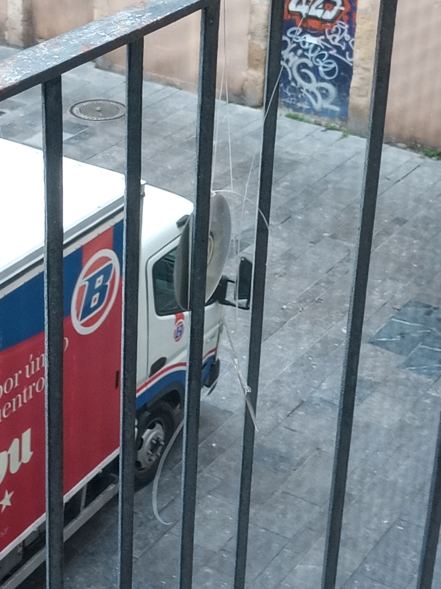 The image shows a street scene in Tarragona, Spain, viewed through a black metal railing. In the foreground, the dark metal bars of the railing create a strong vertical visual element, partially obscuring the scene beyond.
On the left, a white truck with a red and blue side is parked. The side of the truck features a large blue and red circular logo with a white "B" inside, and text that is partially visible, reading "por un" and "entro". The truck has a visible front tire.
The ground appears to be paved with square tiles. In the background, a building wall is visible with graffiti featuring red, white, and blue spray paint. A manhole cover is visible on the tiled ground to the left of the truck. A thin, white, string-like object hangs down from the top of the frame, across the scene. The lighting suggests it is daytime, possibly overcast.