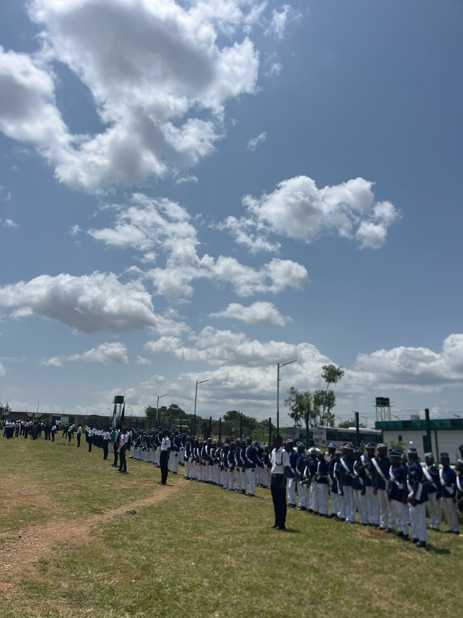 The image depicts a large group of young people, likely cadets or students, standing in formation on a grassy field. They are dressed in matching uniforms consisting of dark blue jackets with white trim, white trousers, and caps. The formation stretches across the frame from left to right and recedes into the distance. A few individuals in similar uniforms are positioned in front of the main group, suggesting some sort of inspection or ceremony. In the background, there are various structures including buildings, a water tower, and fences, indicative of an institutional setting. The sky is partly cloudy with bright sunlight. The scene appears to be outdoors in a relatively open area, with hints of trees and other vegetation around the perimeter. The location is identified as Barikin Jirga Summa, Nigeria.
