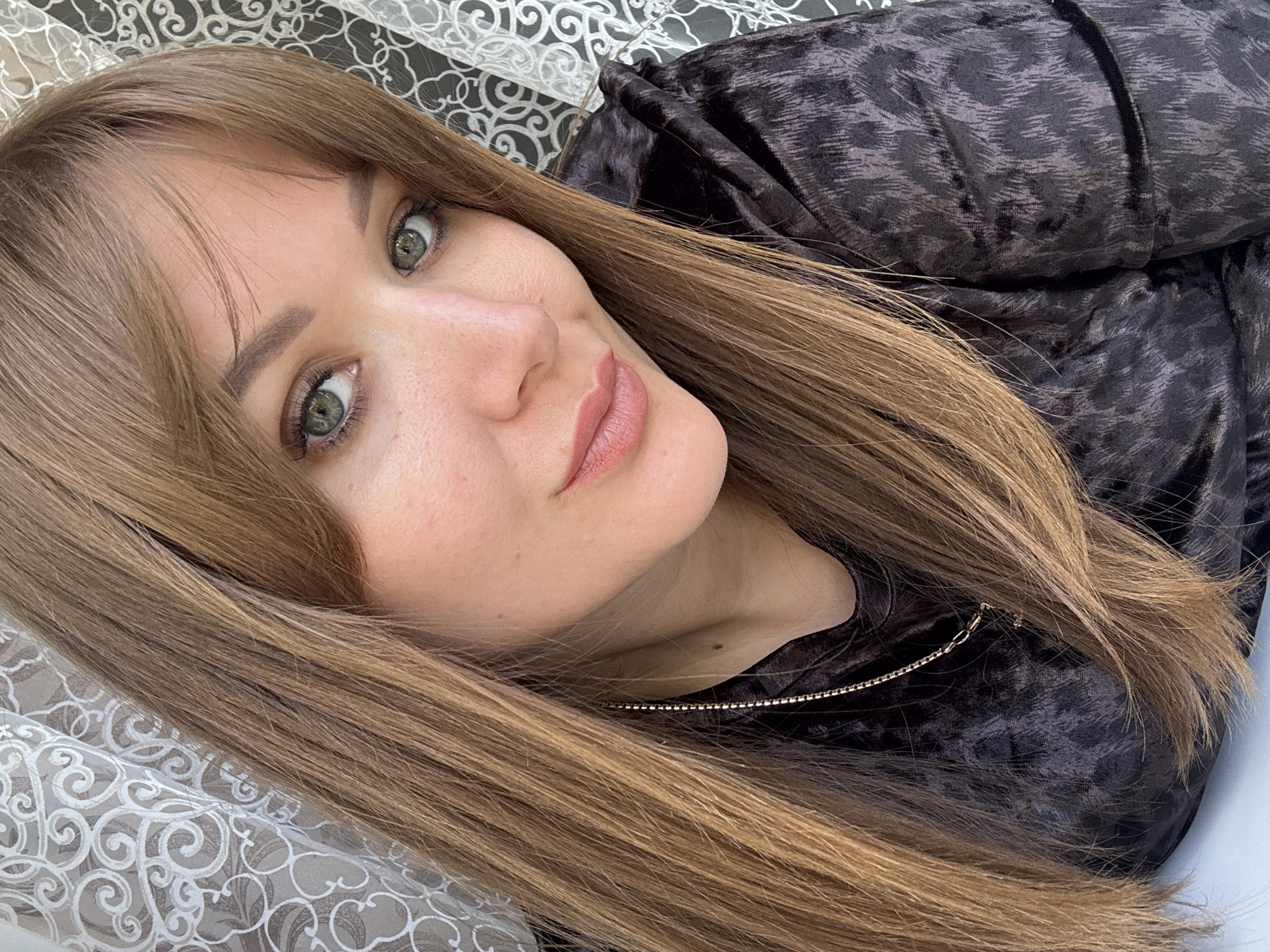 A woman is shown lying down, her head resting on a lacy, patterned surface. She has long, straight, light brown hair with bangs, green eyes, and fair skin. She is wearing a dark-colored garment with a subtle patterned texture and a visible metallic zipper detail. The background appears to be a patterned fabric or surface, possibly a curtain or bedding. There is no information available regarding the location of this image in Chaikovsky, Russia, or any city in Russia.
