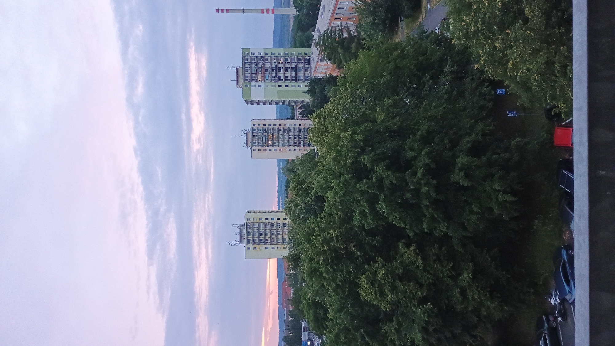 The image shows a wide shot of a residential area in Sokolov, Czechia, at dusk. In the background, three tall apartment buildings are visible, two of which are cream-colored, and one is light green. A red and white striped chimney stack is also visible in the distance to the right of the buildings. In the foreground, a dense canopy of green trees dominates the scene, obscuring much of the lower portion of the apartment buildings. Below the trees, a parking lot with several cars is visible. A row of buildings with orange and white facades can be seen in the middle ground to the right. The sky is a soft pastel hue, transitioning from pale pink at the horizon to a light purple and blue at the top, with wispy clouds scattered across it. The overall impression is one of a quiet, urban evening scene.