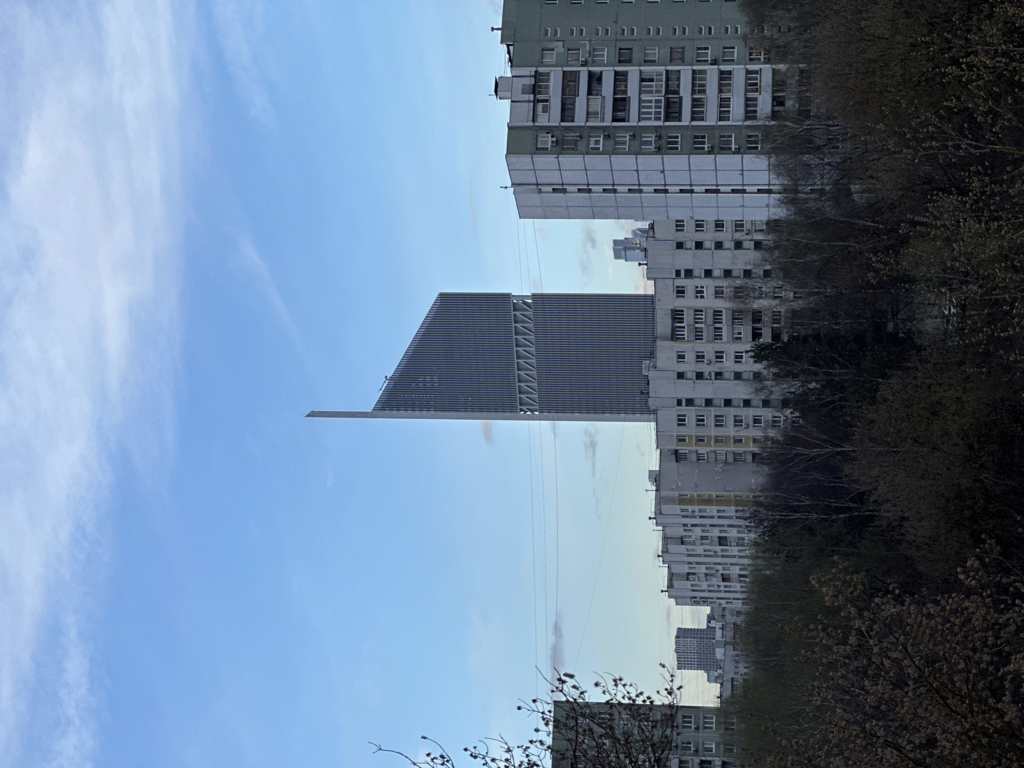 The image shows a view of Khimki, Russia, with a tall, modern skyscraper standing prominently among older, Soviet-era apartment buildings. The skyscraper has a silver exterior with a distinctive angled roof and visible structural supports. The apartment buildings are multi-story, with repetitive rows of windows, and are in various states of repair. In the foreground, there are trees with budding leaves, partially obscuring some of the buildings. The sky is blue with scattered white clouds. Overhead power lines stretch across the scene.