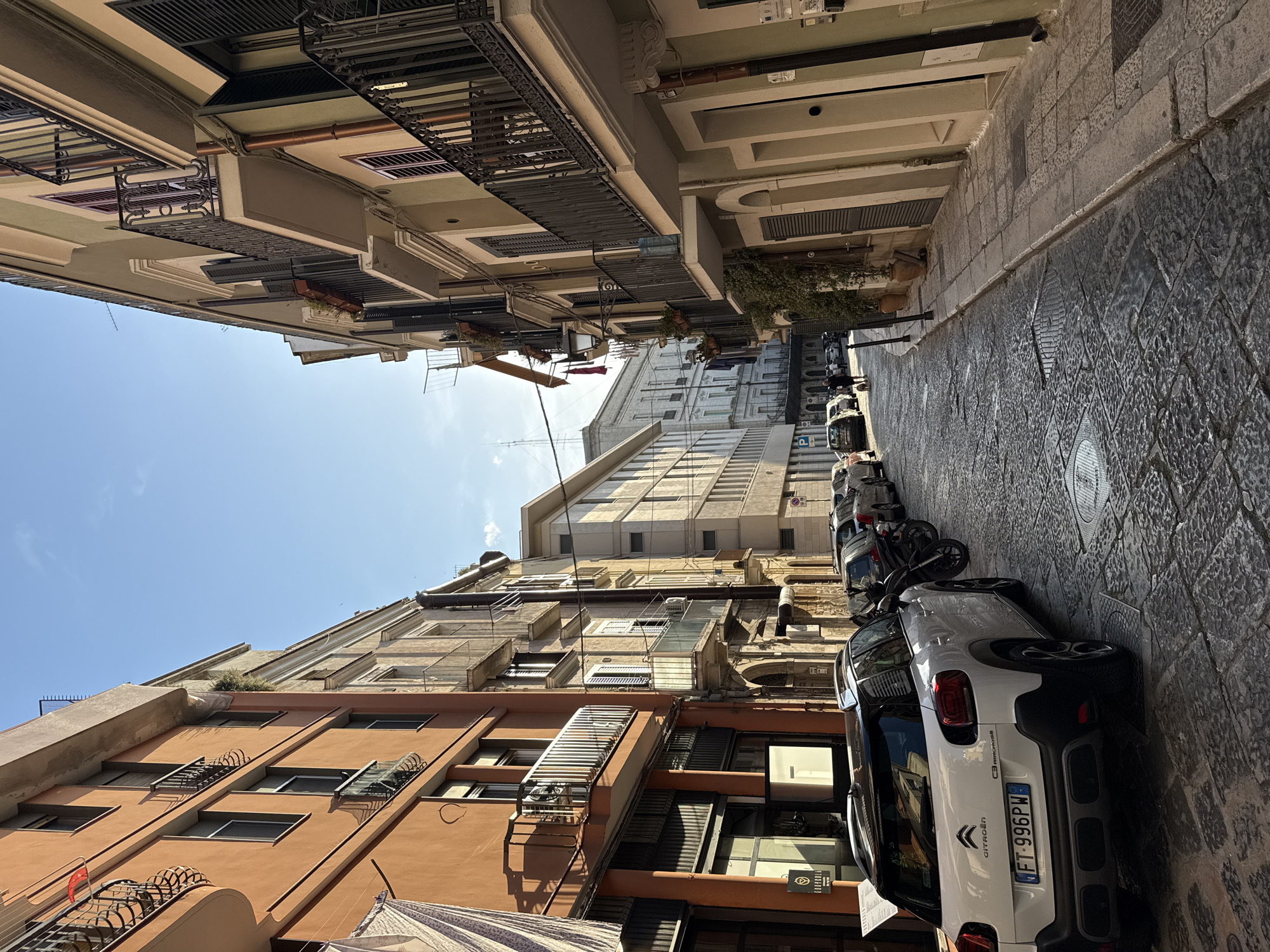 The image displays a sunlit, narrow urban street lined with multi-story buildings, captured from a low perspective looking upwards along the thoroughfare. On the left, a row of residential or commercial buildings rises steeply, characterized by various architectural styles, multiple windows, some with external metal shutters, and balconies adorned with decorative wrought-iron railings. Facades vary from terracotta to lighter cream and beige, with some visible pipes and conduits running along their exteriors.
The street, paved with cobblestones or large irregular stone slabs, runs through the center-right of the frame. Along its right side, several vehicles are parked. Most prominently in the foreground is the rear of a white Citroën C3 Aircross, clearly displaying its "CITROËN" and "C3 AIRCROSS" badging, and a European license plate reading "FT-996PW". Further down the street, other parked cars and at least one motorcycle are visible. A blue "P" parking sign is also discernible.
The sky is bright blue and clear, indicating a sunny day. Strong sunlight creates distinct shadows on the buildings and pavement, emphasizing textures and architectural details. No people are present in the scene. A partially visible shop sign on a building on the left appears to say "EUROSPORTS" or similar. The overall setting is a quiet, densely built street in what appears to be a European city.