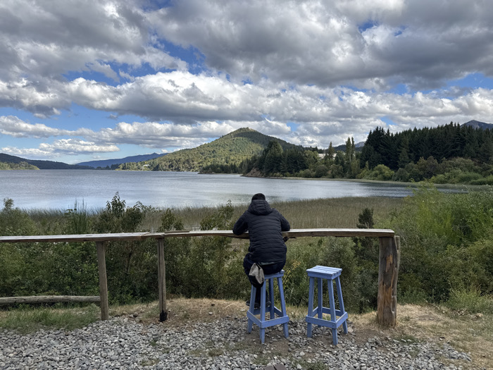 A solitary figure sits on a blue stool, back to the viewer, at a wooden counter overlooking a serene lake. A second stool stands beside them. The landscape includes rolling hills, dense green trees, and reeds along the lake's edge. The sky is filled with dramatic clouds, casting diffused light on the scene. The foreground consists of a gravelly area, enhancing the sense of being in a tranquil, natural setting.
