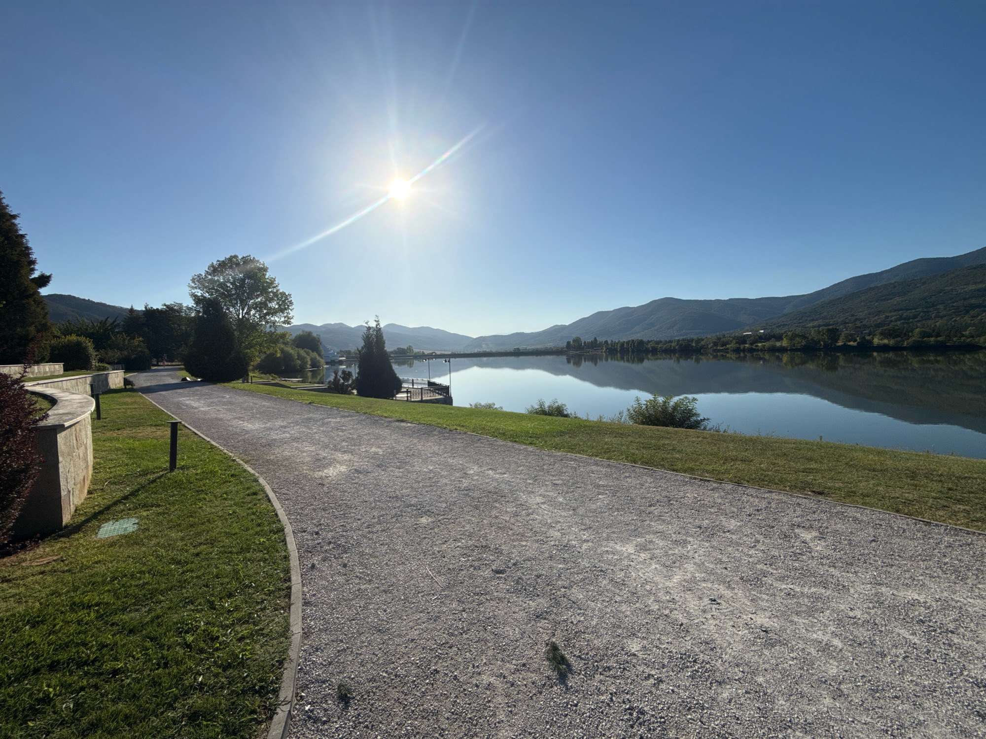 The image shows a scenic view of a lake and surrounding mountains on a clear, sunny day. In the foreground, a gravel path curves to the right, bordered by a neatly mown lawn. A low, curved stone wall with plantings is visible on the left side of the path. In the midground, trees and some structures, including a pier with lights, line the edge of the tranquil lake, which reflects the clear blue sky and the distant, tree-covered hills. The sun is high in the sky, creating a bright glare and lens flare. The location is the lake in the city of Pravets, Bulgaria.