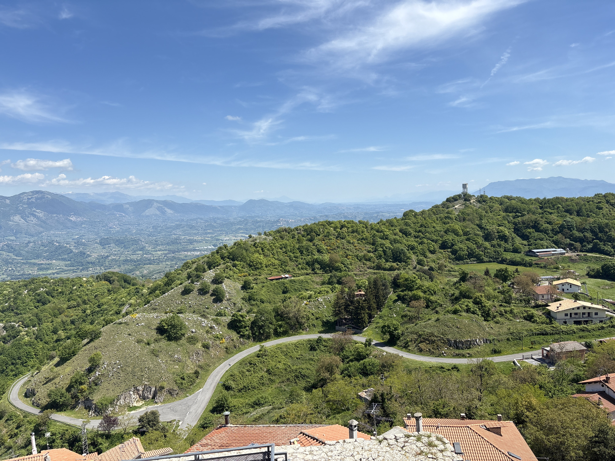 The image shows a landscape view in Rocca di Cave, Italy, with a winding road ascending a green, wooded hillside. In the distance, a valley stretches out, with mountains visible on the horizon beneath a blue sky with scattered clouds. On the hilltop to the right, a tall tower stands amid the trees. In the foreground, the red tile roofs of houses are visible. No people are visible in the frame.