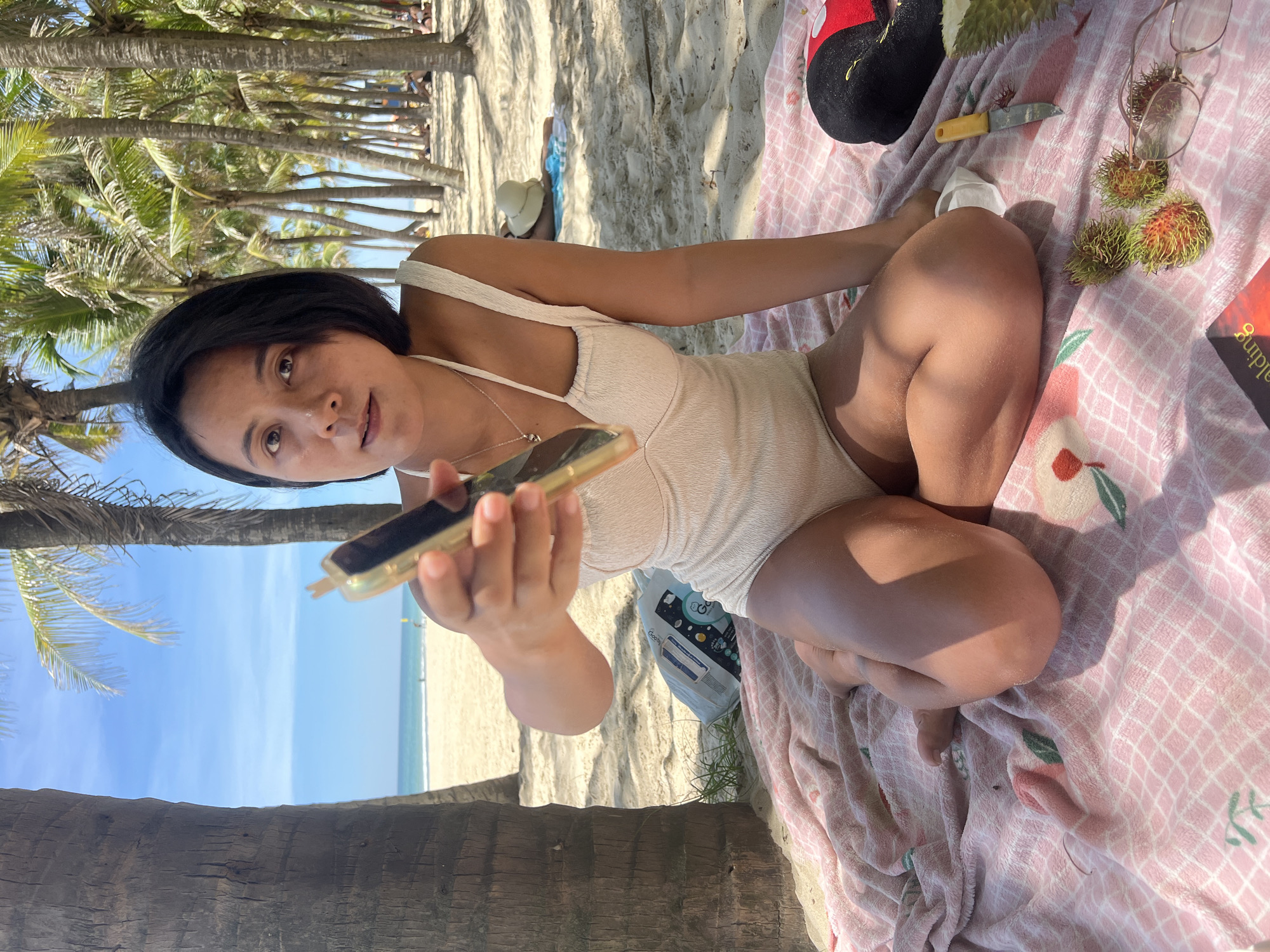 A woman is sitting on a pink and white checkered blanket on a sandy beach, likely Mỹ Khê Beach in Đà Nẵng, Vietnam. She is wearing a light-colored, textured swimsuit. Her legs are crossed in front of her, and she is holding up a smartphone with a gold case towards the camera. In front of her on the blanket are three rambutans, a pair of eyeglasses, a small knife with a yellow handle, and a portion of a durian. Behind her, a large palm tree trunk is visible on the left, and a line of palm trees stretches across the background, with the ocean and a clear blue sky beyond. Other people are visible in the distance on the beach.