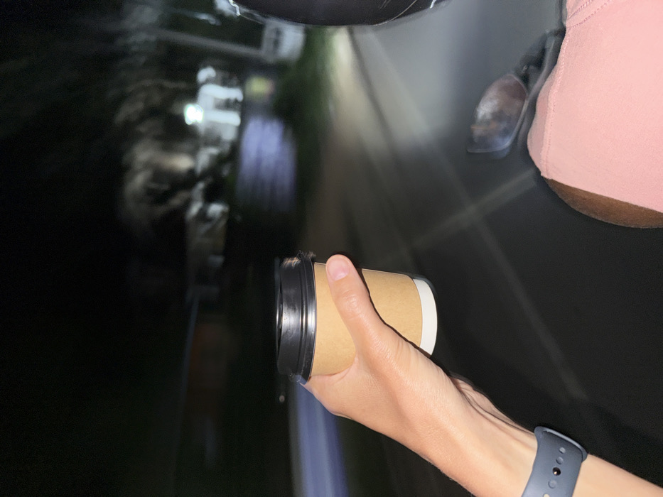 At night in Dikwella, Sri Lanka, a right human hand is depicted holding a standard disposable beverage cup. The cup is light brown with a white lower section and a black plastic lid. A dark blue wristband, consistent with a smartwatch or fitness tracker, is visible on the individual's left wrist in the lower-left corner. A portion of a light pink garment, likely worn by the same individual, is visible on the right side of the frame, below a dark-colored helmet. A blurred metallic object, possibly a vehicle rearview mirror or bodywork, is present below the helmet. The scene is observed from a moving vehicle, indicated by motion blur in the background. The environment is dark, with indistinct, blurred shapes of trees or foliage and distant artificial lights. A single prominent light source illuminates a section of the asphalt road ahead, casting a glare.