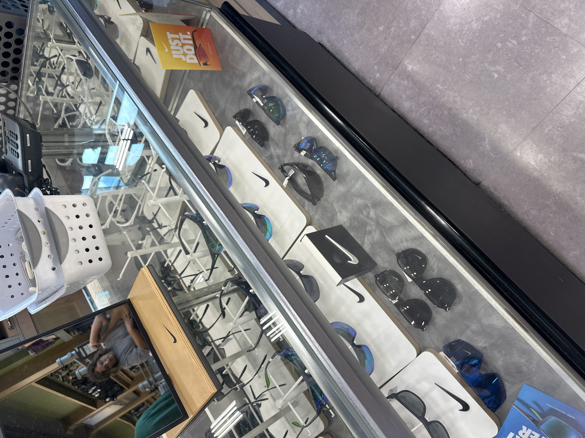 The image shows a retail display case filled with Nike sunglasses. The display case is made of glass and has a black base. The sunglasses are arranged on white and wooden stands, some with Nike logos. There are also some informational displays with the Nike "Just Do It" slogan. In the reflection of the glass, a person with curly hair wearing a gray t-shirt can be seen adjusting their hair. The background appears to be a store environment with shelves and other retail items. The flooring is light gray. The image is taken from a slightly elevated, angled perspective looking down into the display case. The location is Algonquin, United States of America.