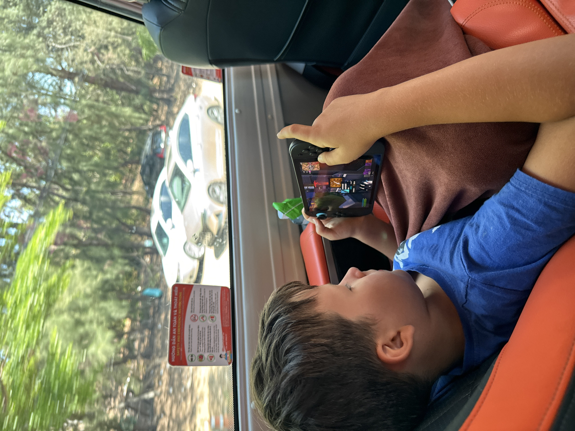 Inside a vehicle, likely a bus or van, a young boy is engrossed in playing a video game on a smartphone with an attached controller. He is wearing a blue t-shirt and is resting against an orange seat and a brown pillow or blanket. His face is visible in profile, showing concentration on the game screen, which displays a colorful, intricate interface.
Through the vehicle's window, an outdoor scene unfolds under bright daylight. Tall, slender trees with green foliage line what appears to be a road or parking area. Several cars, predominantly white and dark-colored, are visible outside. The weather appears sunny and clear.
Affixed to the window frame is a red and white safety instruction card. The visible text on this card includes "HƯỚNG DẪN AN TOÀN VÀ THOÁT HIỂM" and its English translation, "SAFETY AND EMERGENCY INSTRUCTIONS." The setting for this scene is Mũi Né, Vietnam. The image captures a moment of quiet entertainment during travel.